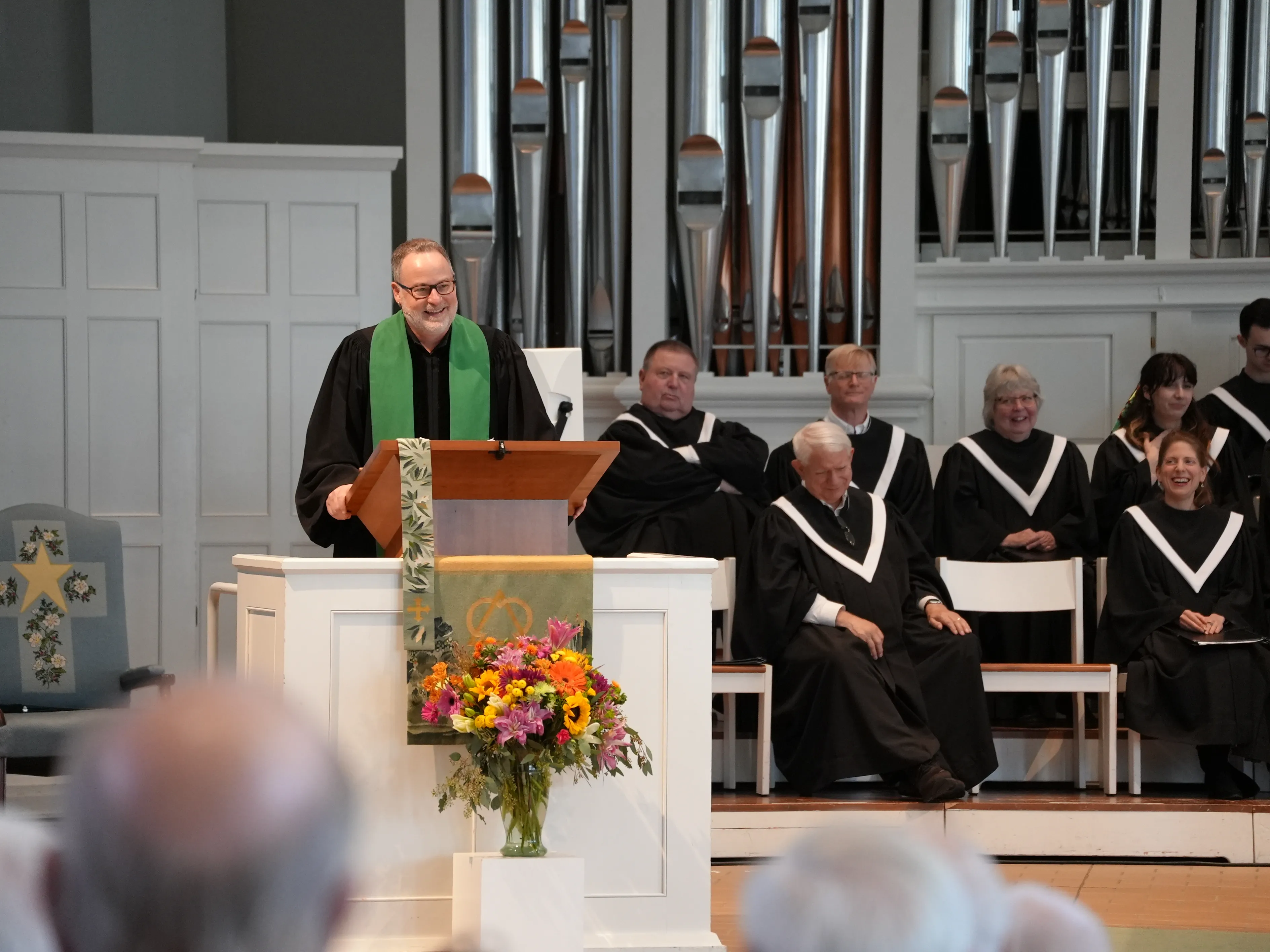 Clergyman with a green stole speaking at a church pulpit decorated with flowers, with choir members seated behind.