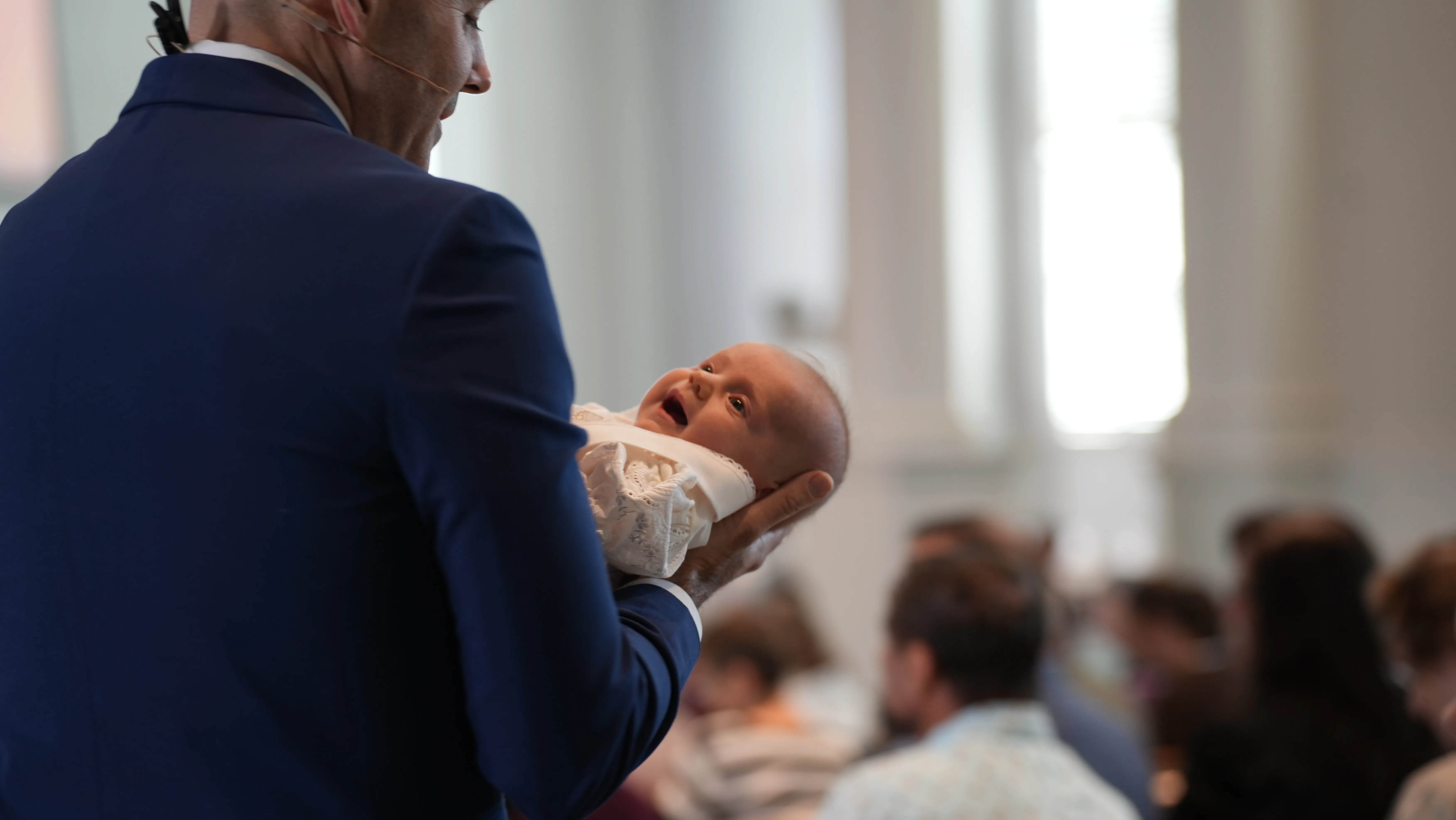 Man in blue suit holding a smiling baby dressed in white in a room with an audience.