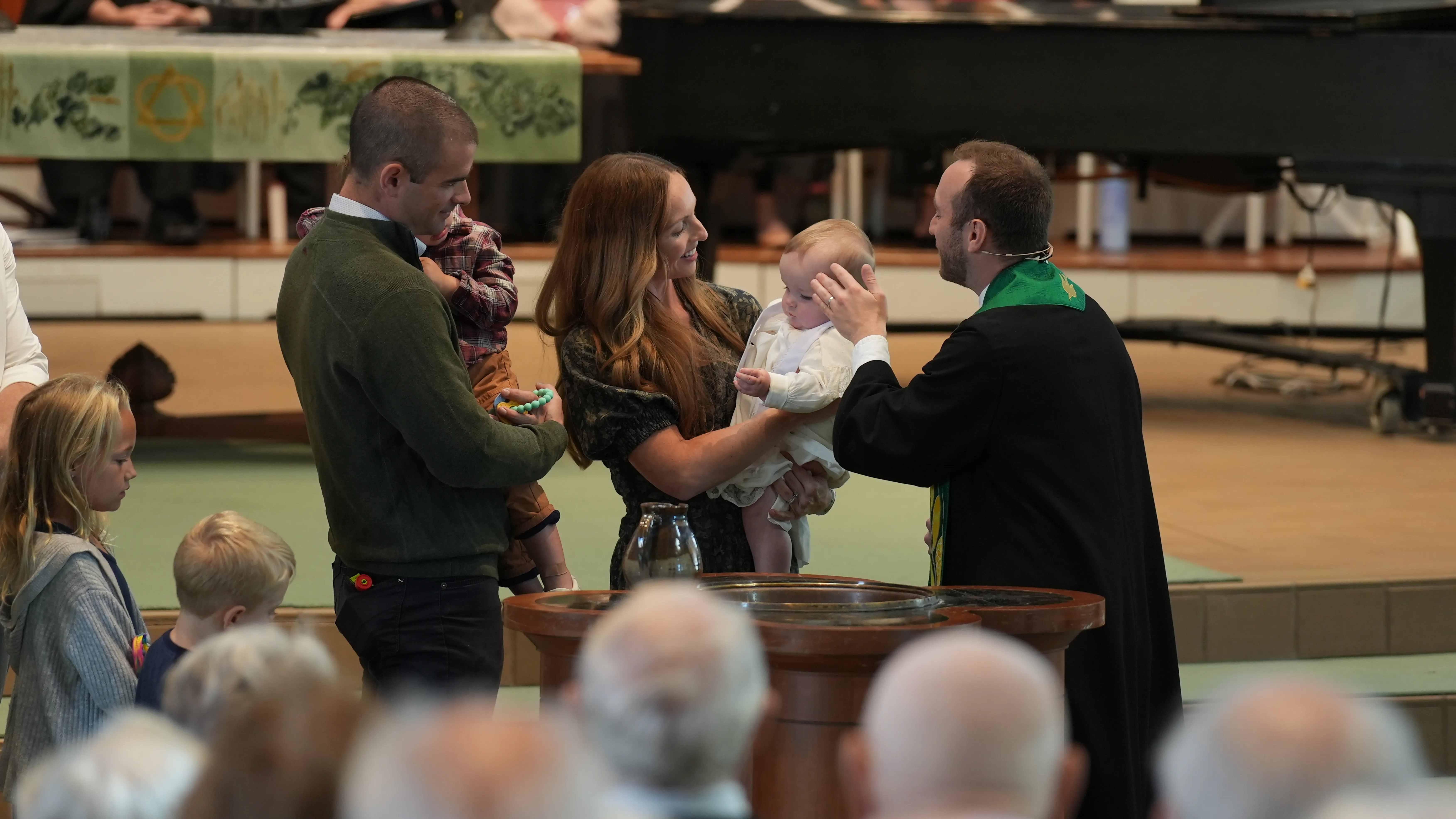 A priest in black robe and green stole blessing a baby held by a smiling woman during a baptism ceremony with family nearby.