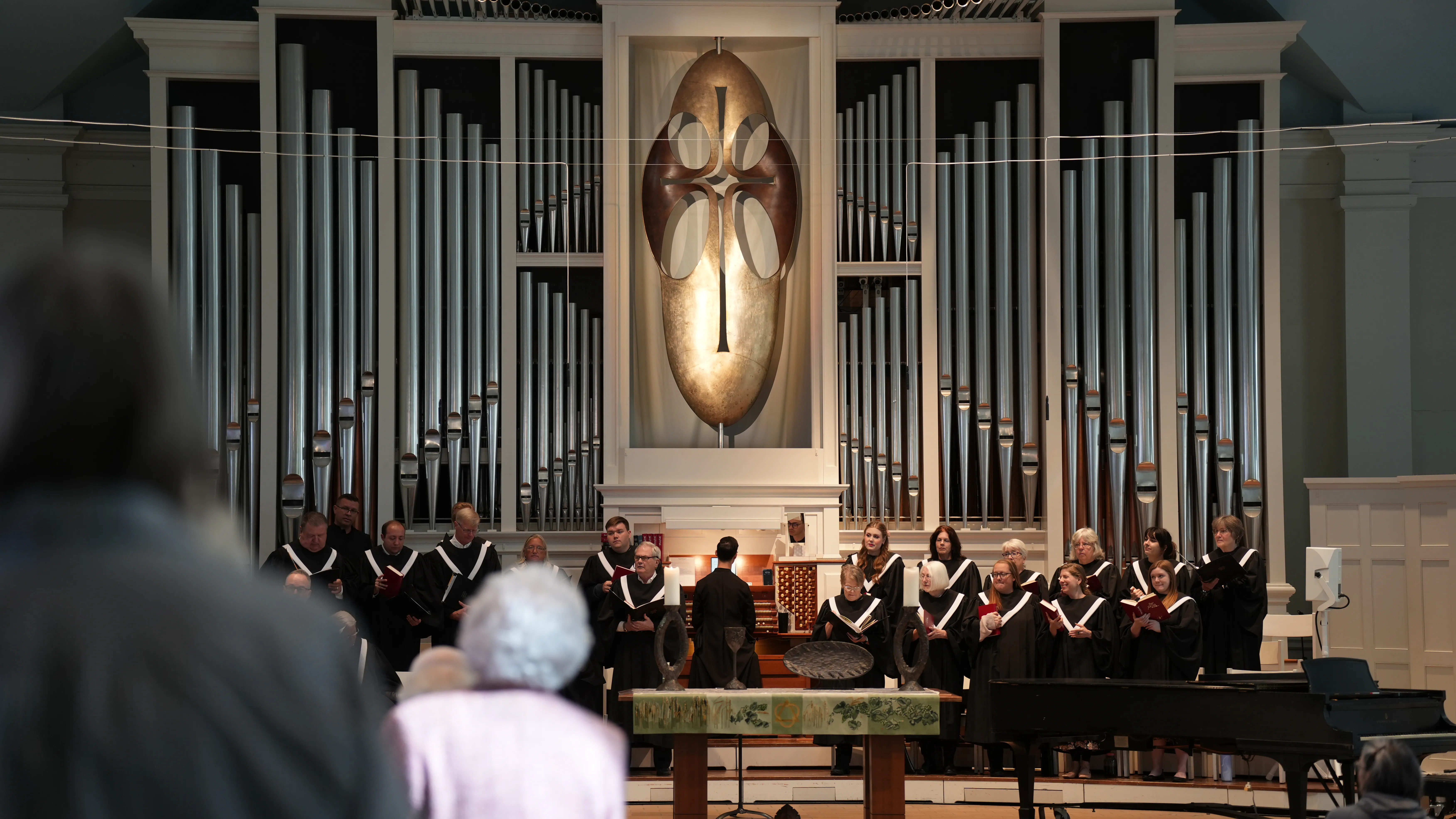 Church choir dressed in black robes with white collars singing in front of a large pipe organ and a cross-shaped wall decoration.