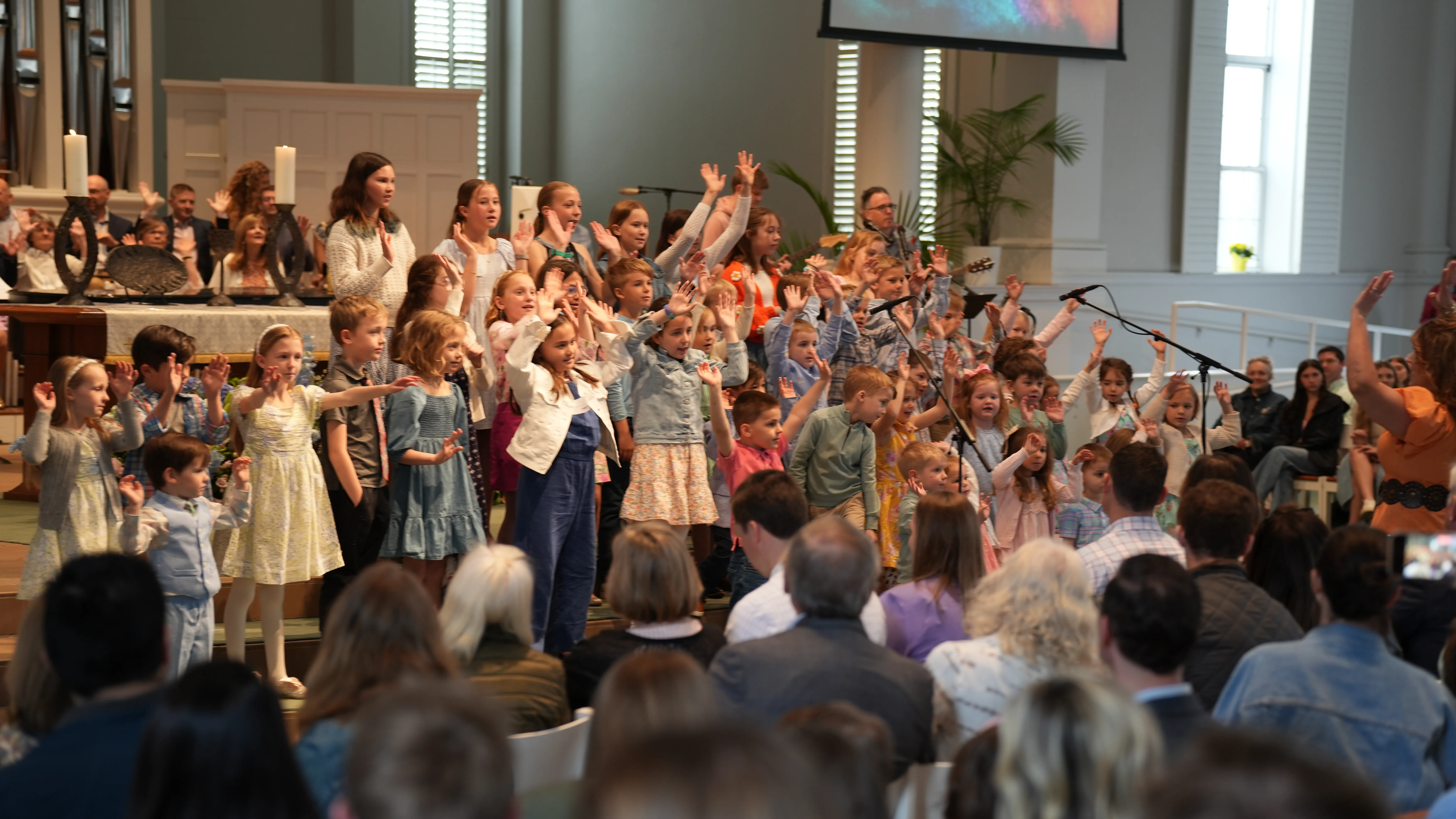 Large group of children standing on a church stage singing and raising hands, directed by a woman in orange.