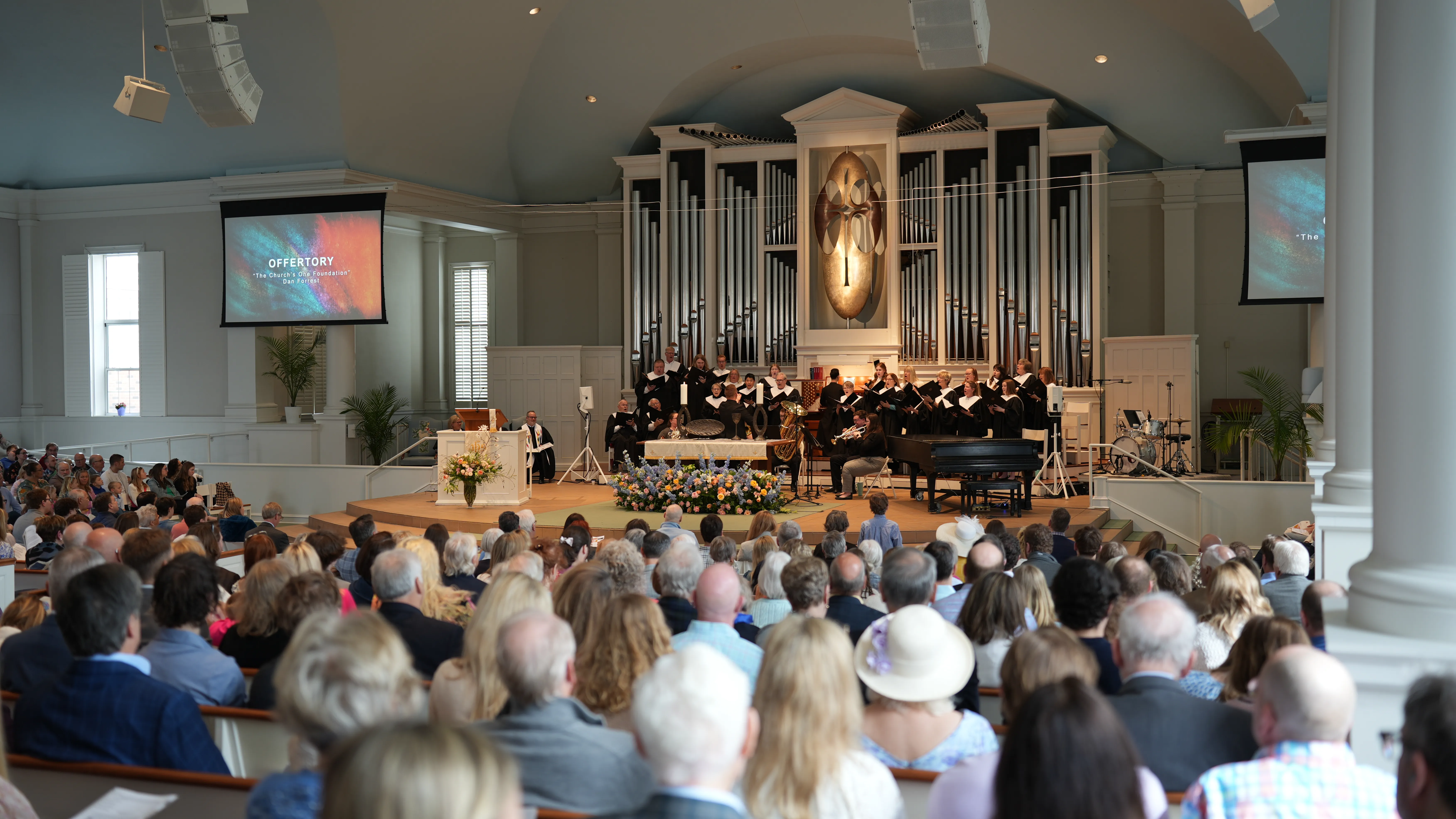 Congregation attending a church service with choir and musicians performing on stage in front of a large pipe organ.