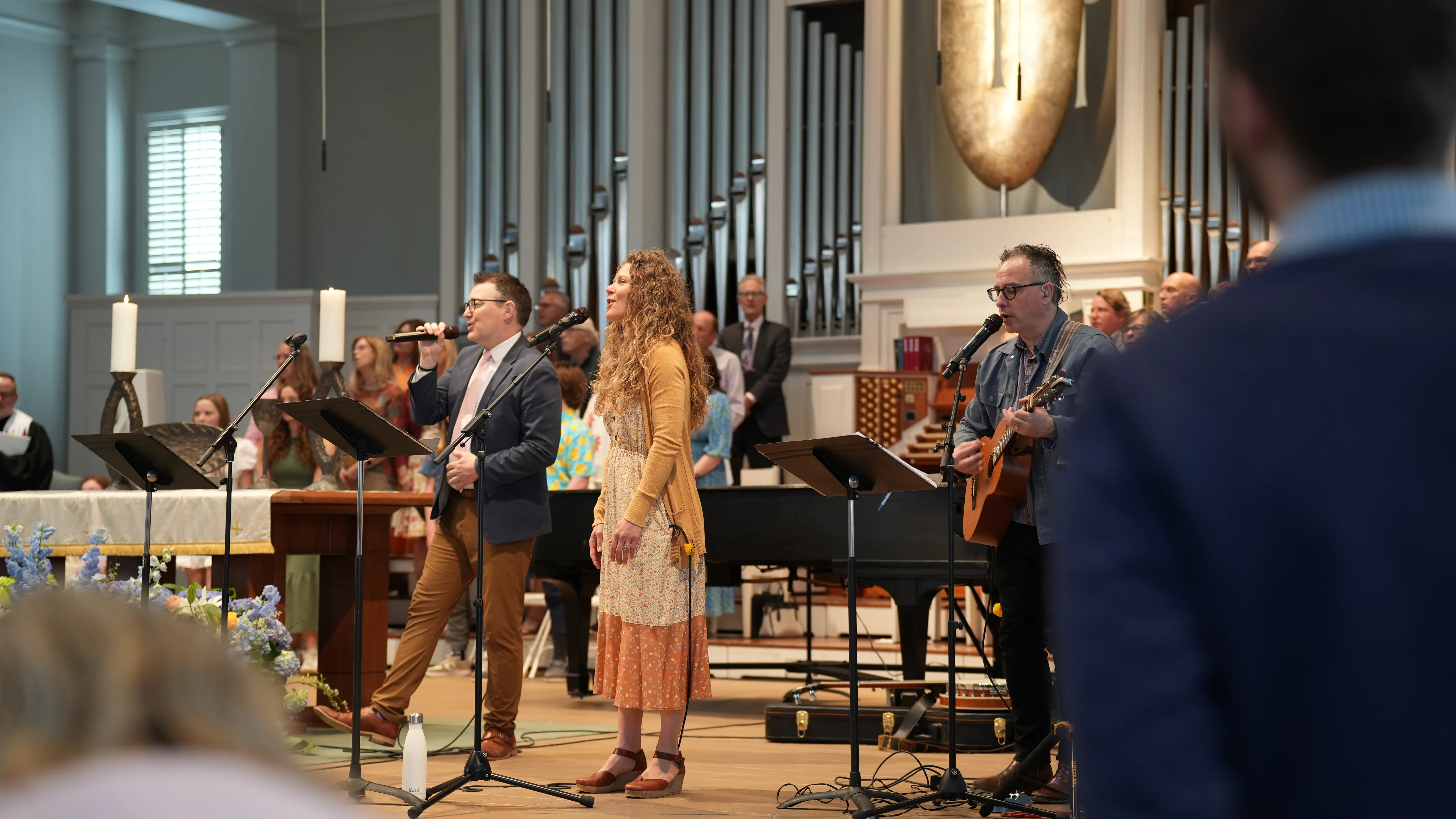 Three musicians performing with microphones and a guitar in a church with an organ in the background.