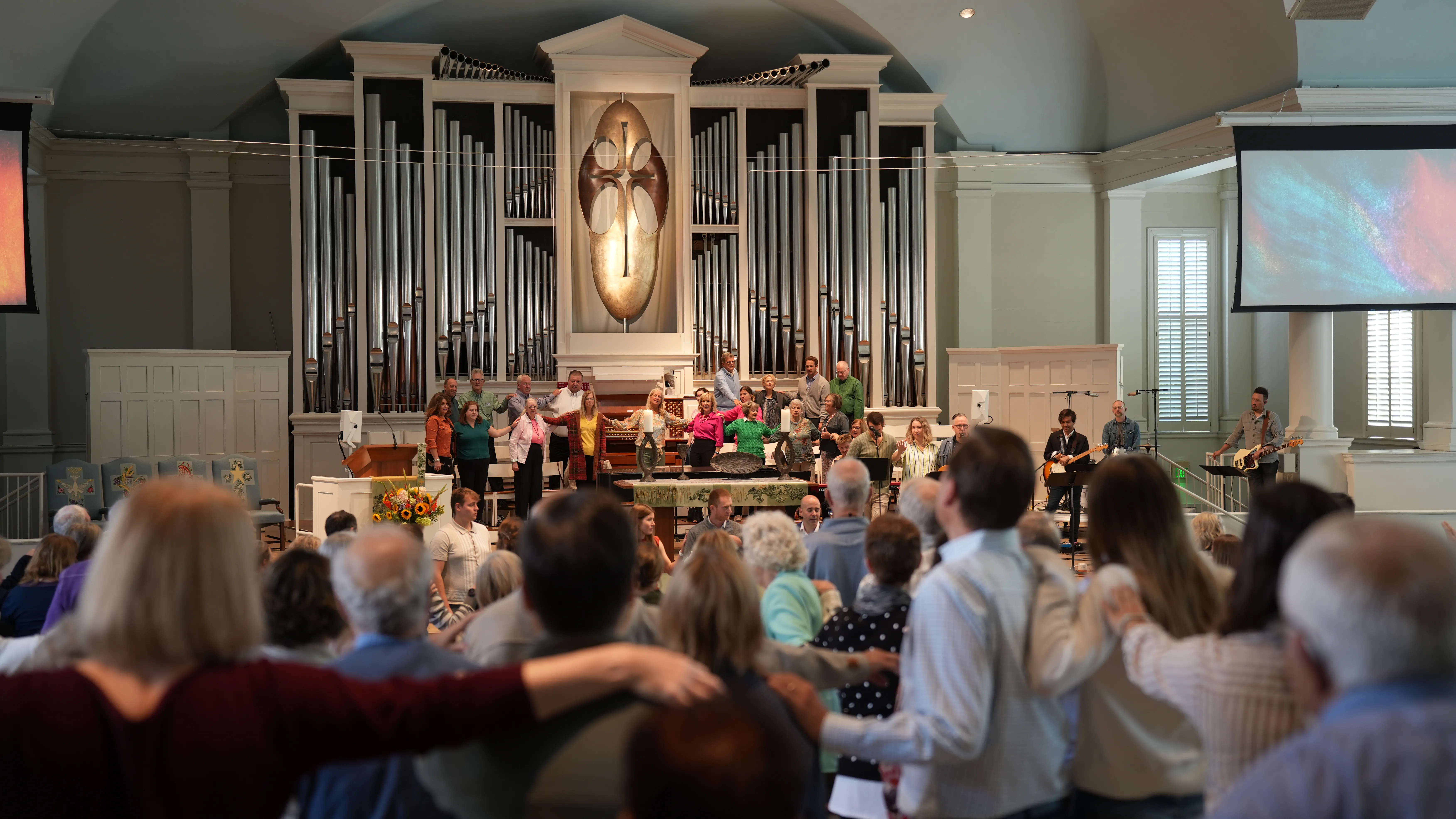 Congregation standing and holding hands toward a choir and musicians in a church with a large pipe organ and cross emblem.