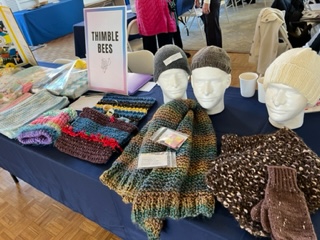 Table display of colorful knitted hats, scarves, and gloves with a sign reading 'Thimble Bees' at a craft fair.