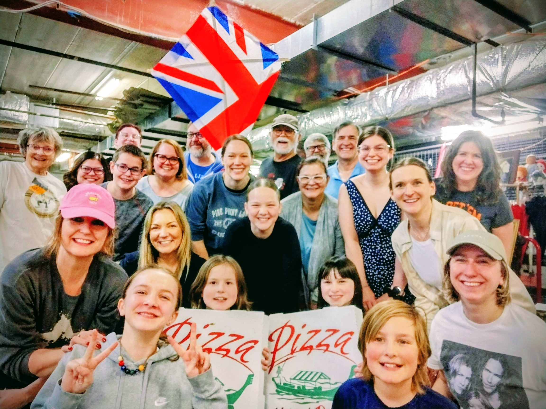 Group of smiling people of various ages posing indoors with two pizza boxes and a British flag in the background.