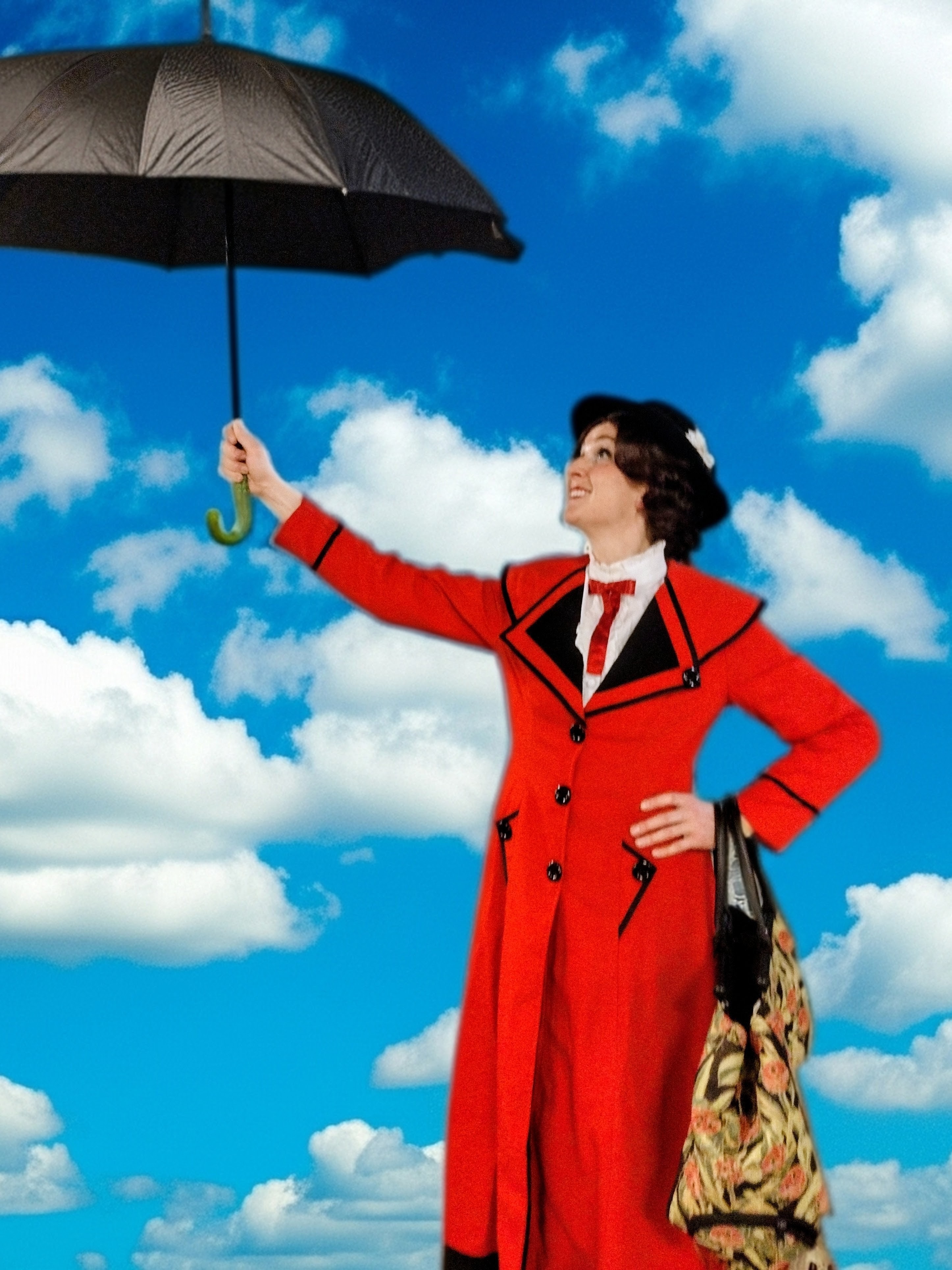 Woman in a red coat and black hat holding a black umbrella against a bright blue sky with white clouds.