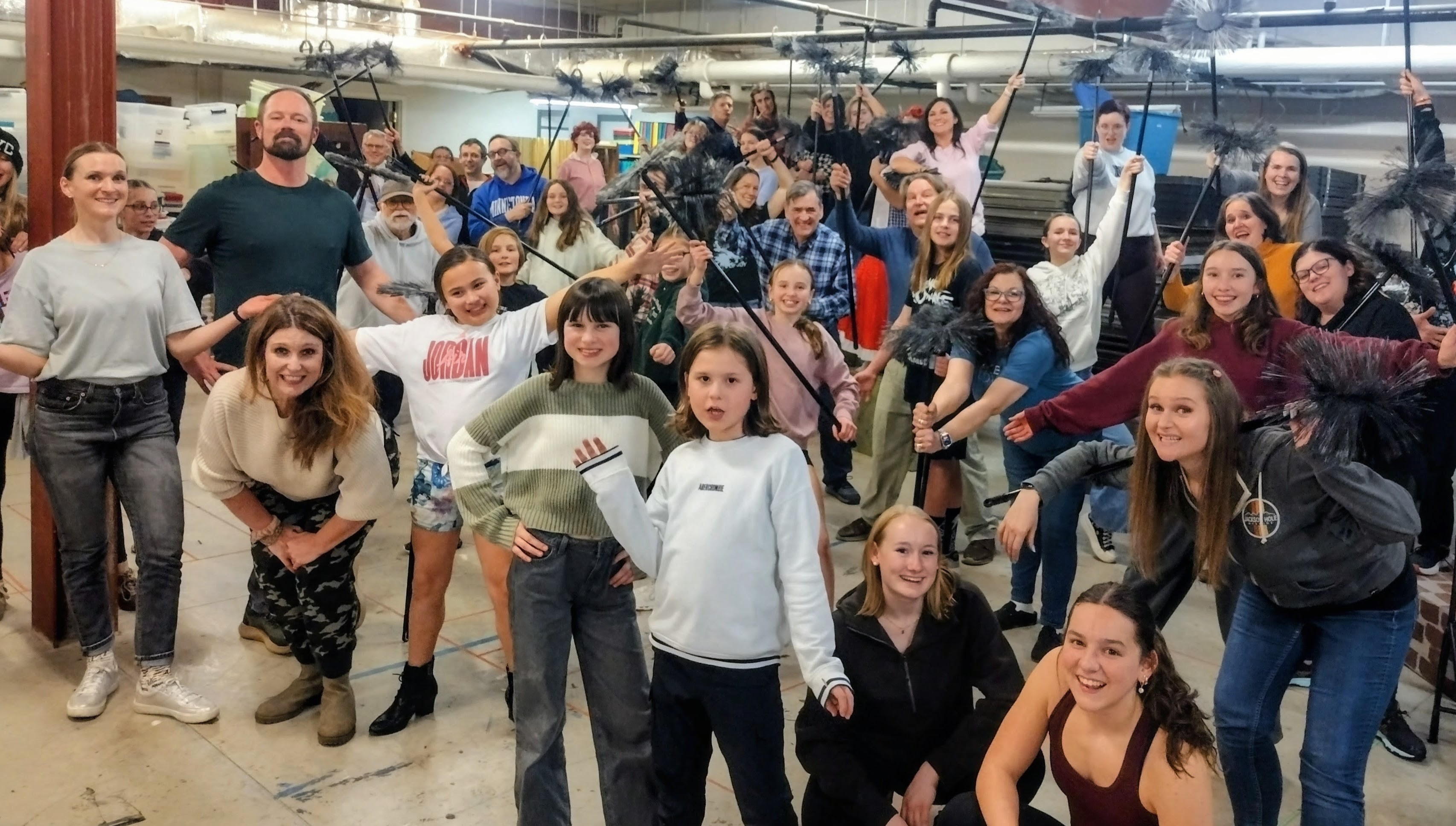Large group of people of various ages gathered indoors, many holding chimney sweeping brushes raised in the air, smiling and posing for the photo.