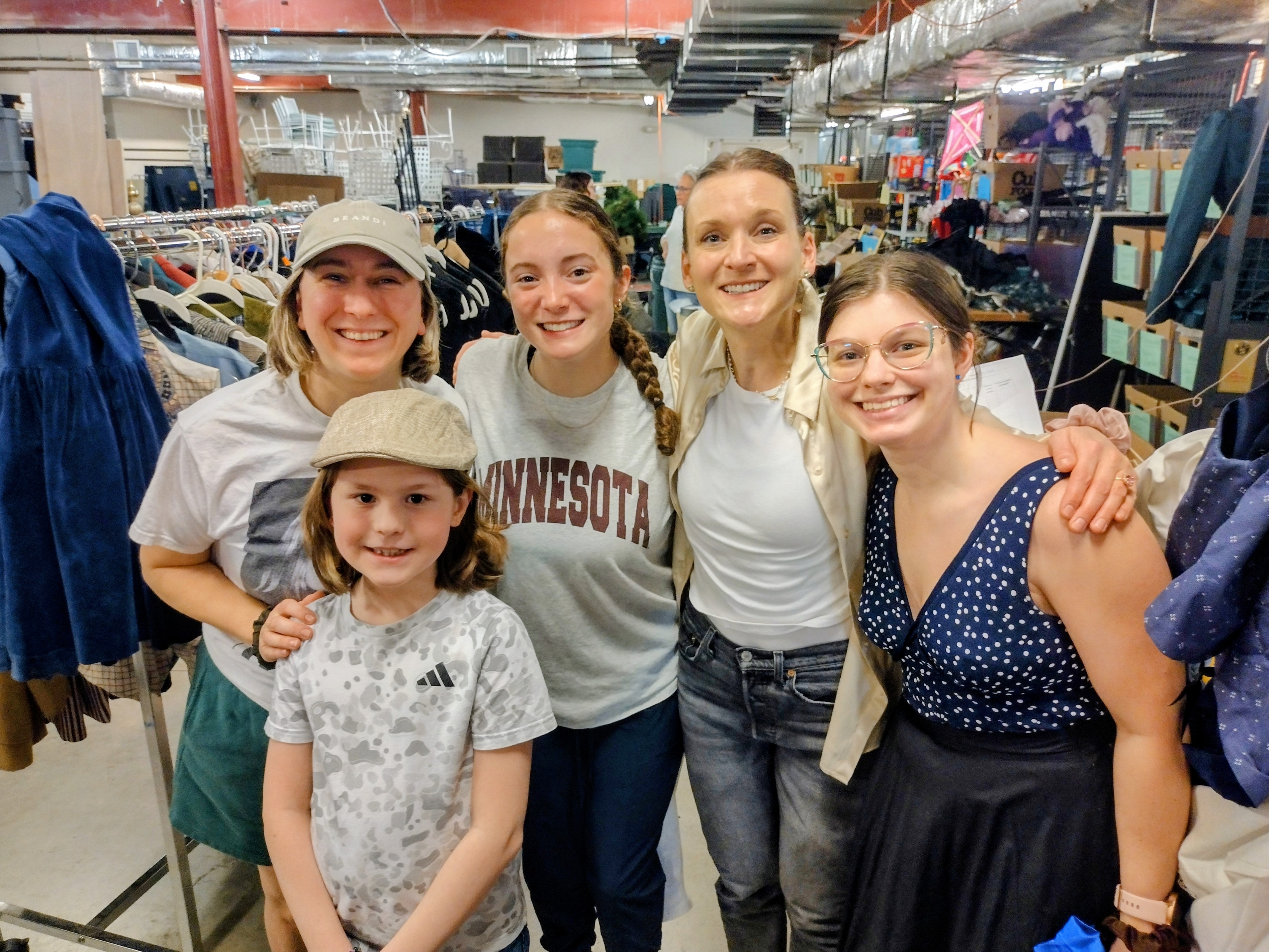 Five smiling people posing together in a warehouse with clothing racks and shelves in the background.