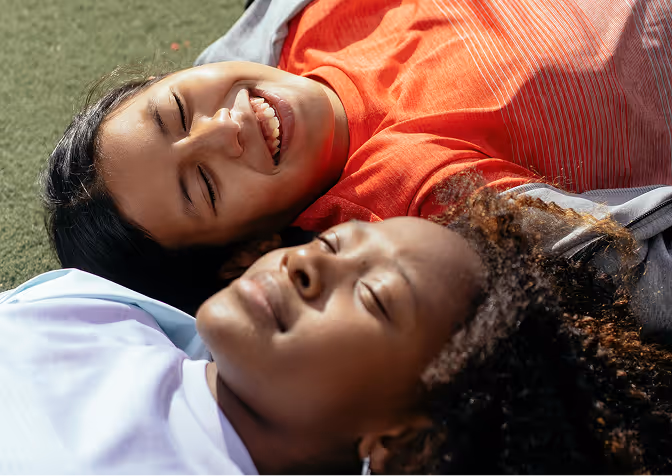 Two young women lying on grass, one smiling with eyes closed, the other relaxed with eyes closed.