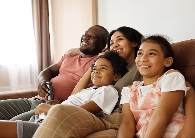 Smiling family of four sitting closely together on a couch watching television, with father holding a remote control.