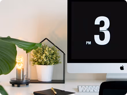 Modern workspace with an iMac displaying a digital clock showing 3 PM, a white potted plant, a lit decorative bulb inside a glass container, a notebook with a pen, and a keyboard on a white desk.