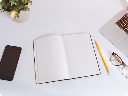 White desk with a blank open notebook, yellow pencil, eyeglasses, smartphone, laptop, and a small potted plant.