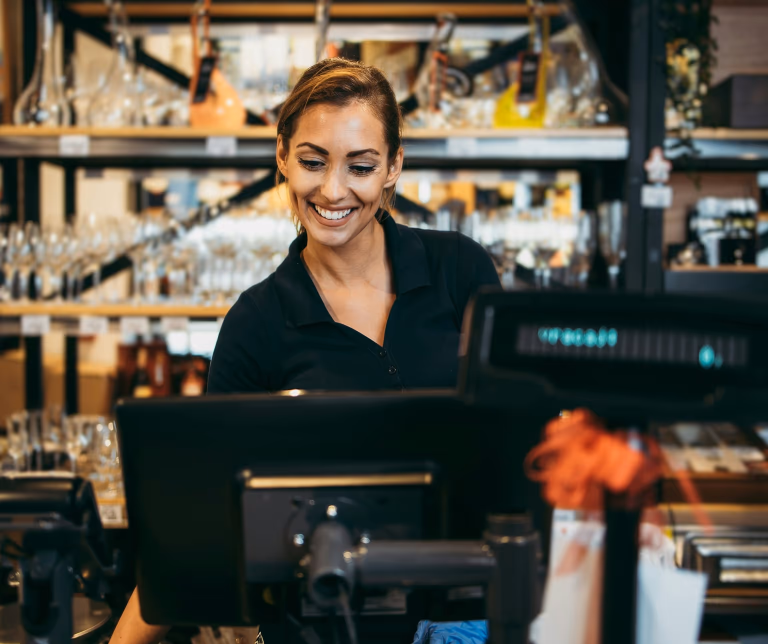 Serveuse souriante regardant un écran de caisse enregistreuse dans un bar ou restaurant avec étagères de verres en arrière-plan.