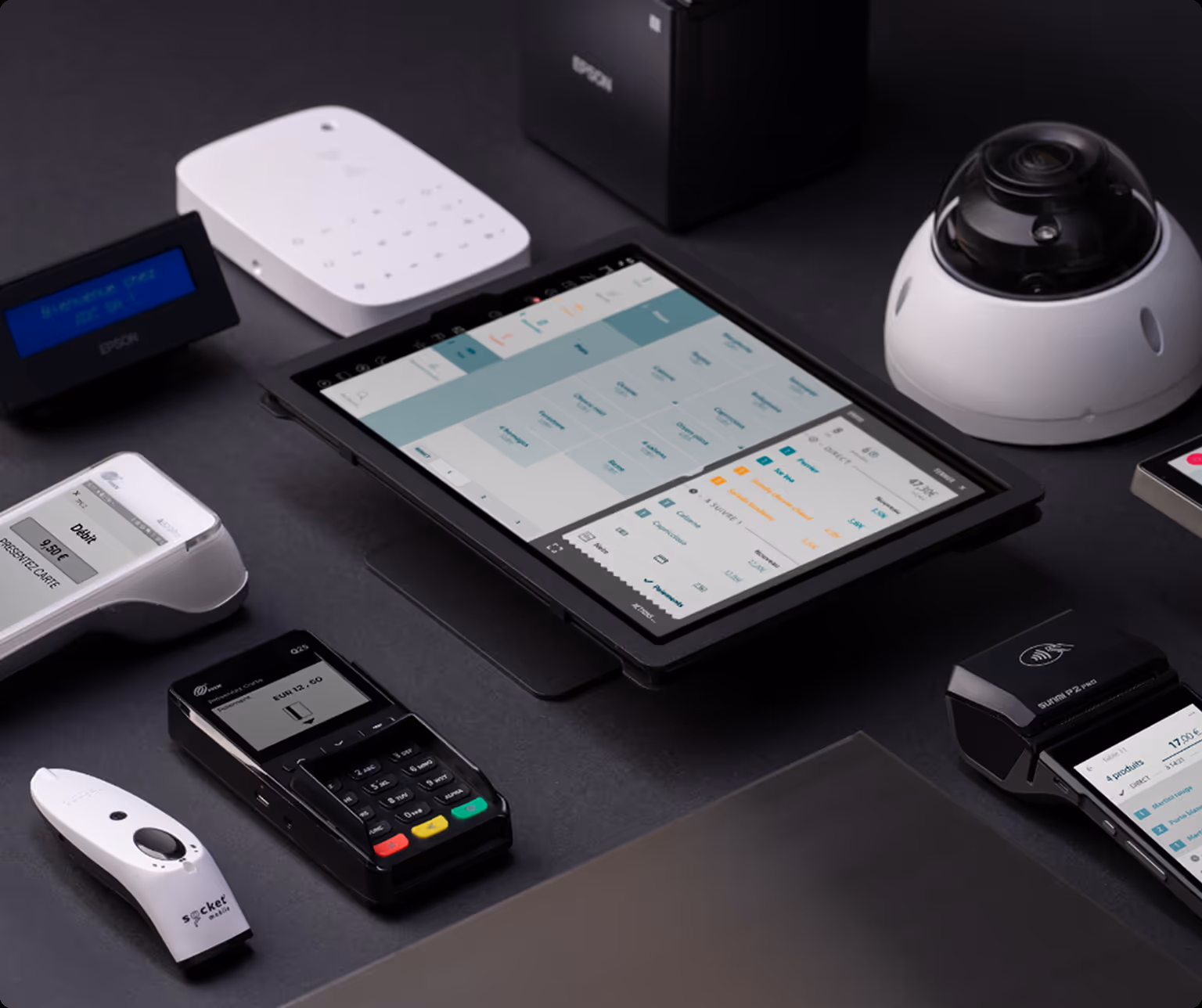 Ensemble d'appareils de point de vente comprenant une tablette affichant un logiciel de caisse, plusieurs terminaux de paiement, un scanner et une caméra de sécurité sur une surface noire.