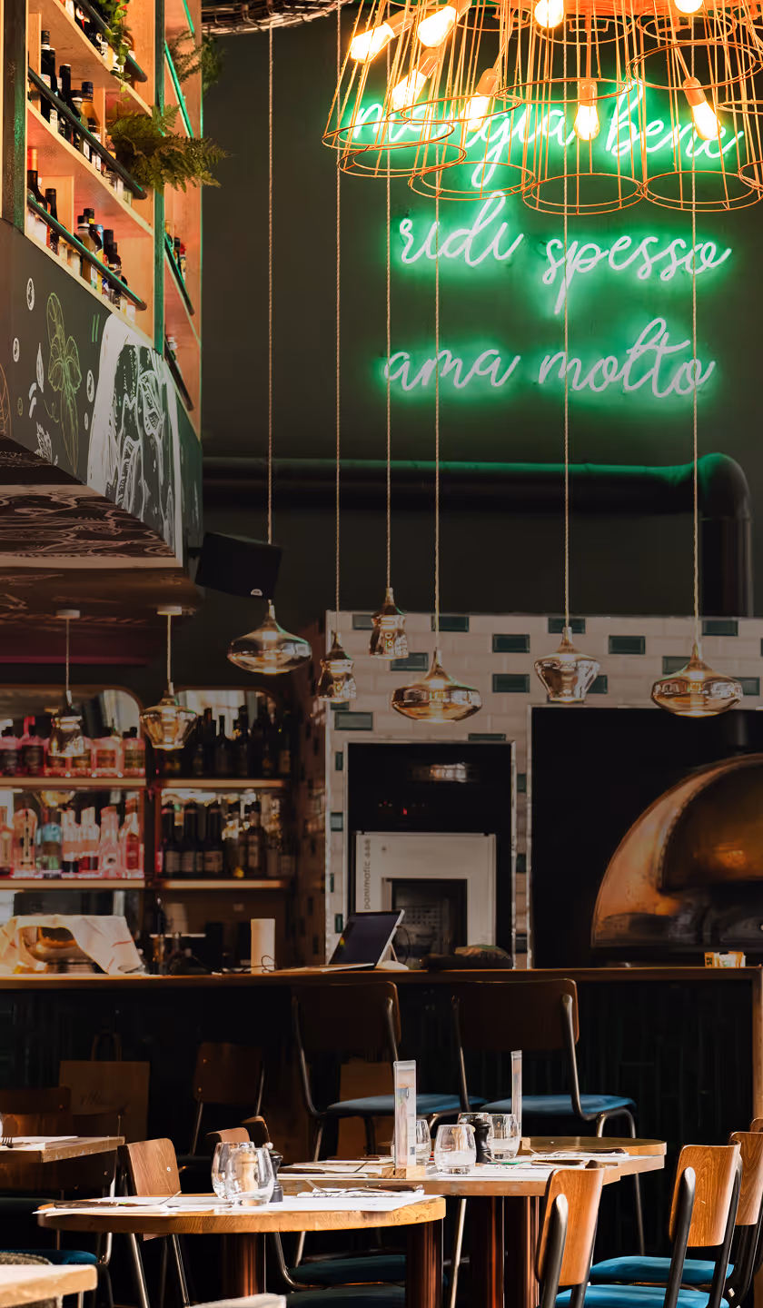 Intérieur d'un restaurant moderne avec tables en bois dressées, chaises, une lampe suspendue et une enseigne lumineuse verte au mur.