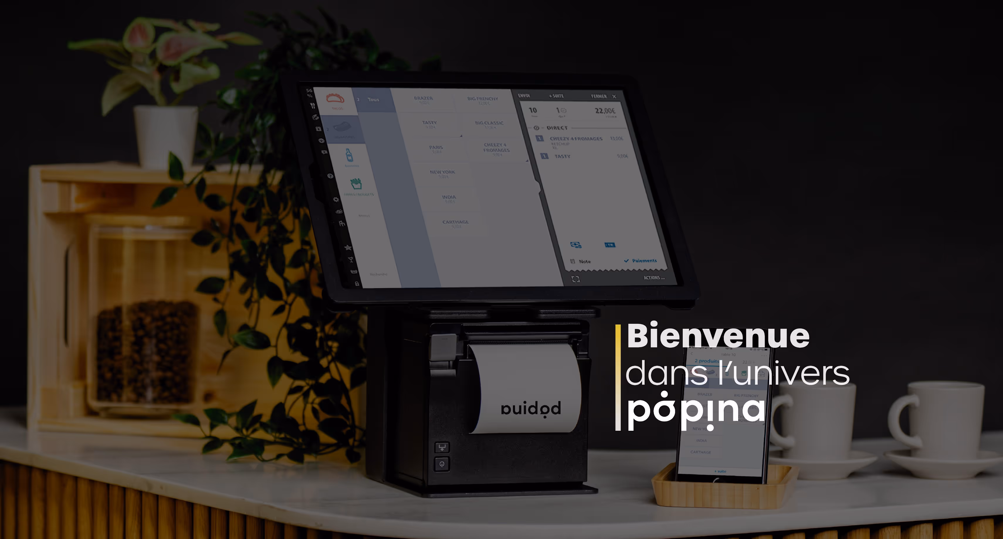 Terminal de point de vente avec écran tactile et imprimante de tickets sur un comptoir, entouré de tasses, plante et accessoires.