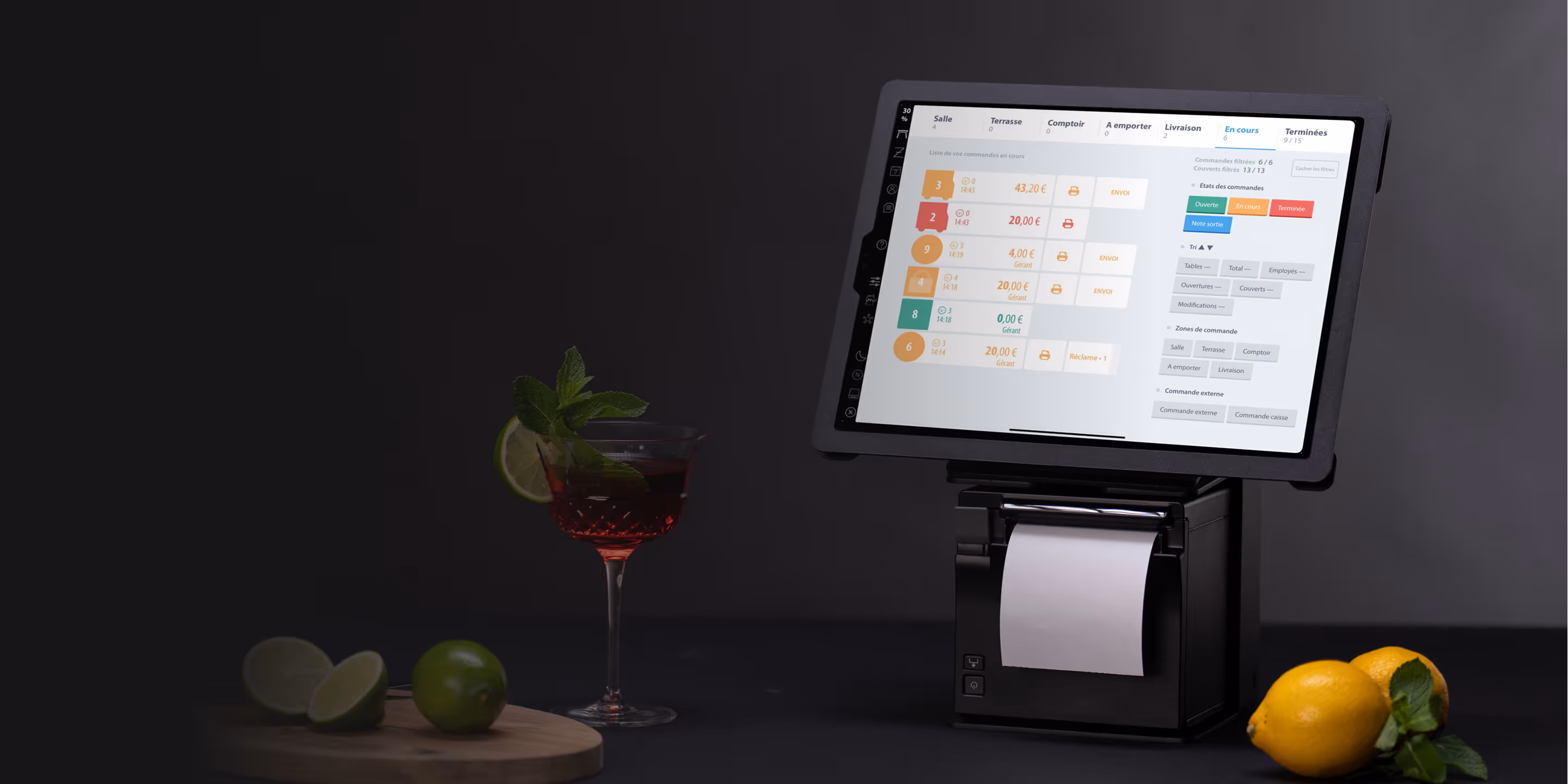 Système de point de vente avec écran tactile affichant des commandes en cours, avec un verre de cocktail, des citrons verts et des citrons jaunes à côté.