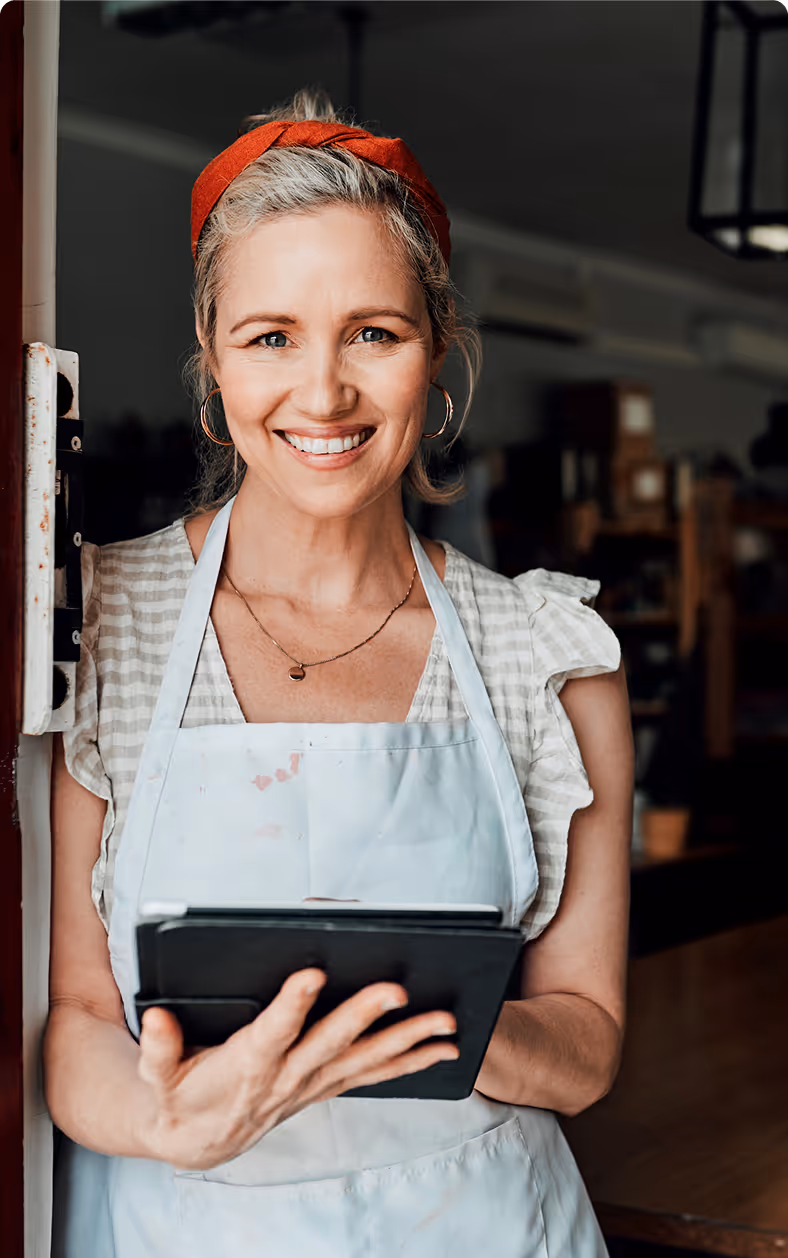 Femme souriante portant un tablier et un bandeau rouge, tenant une tablette numérique en intérieur.