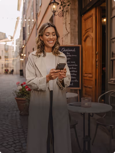 Femme souriante en manteau beige regardant son téléphone dans une rue pavée avec une table et des chaises de café. Elle vient de recevoir une offre marketing par SMS.