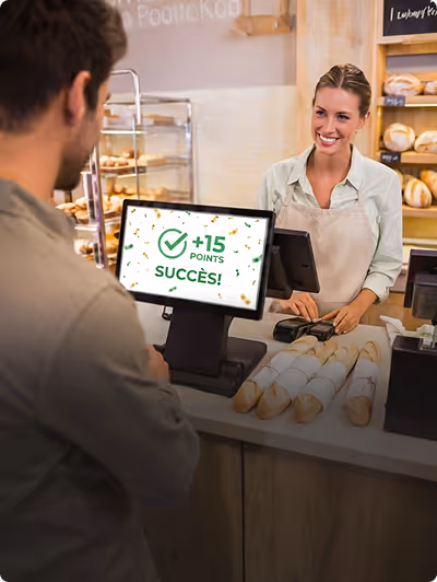 Une vendeuse souriante dans une boulangerie remet un reçu à un client devant un écran affichant '+15 points succès' grâce à son programme de fidélité.