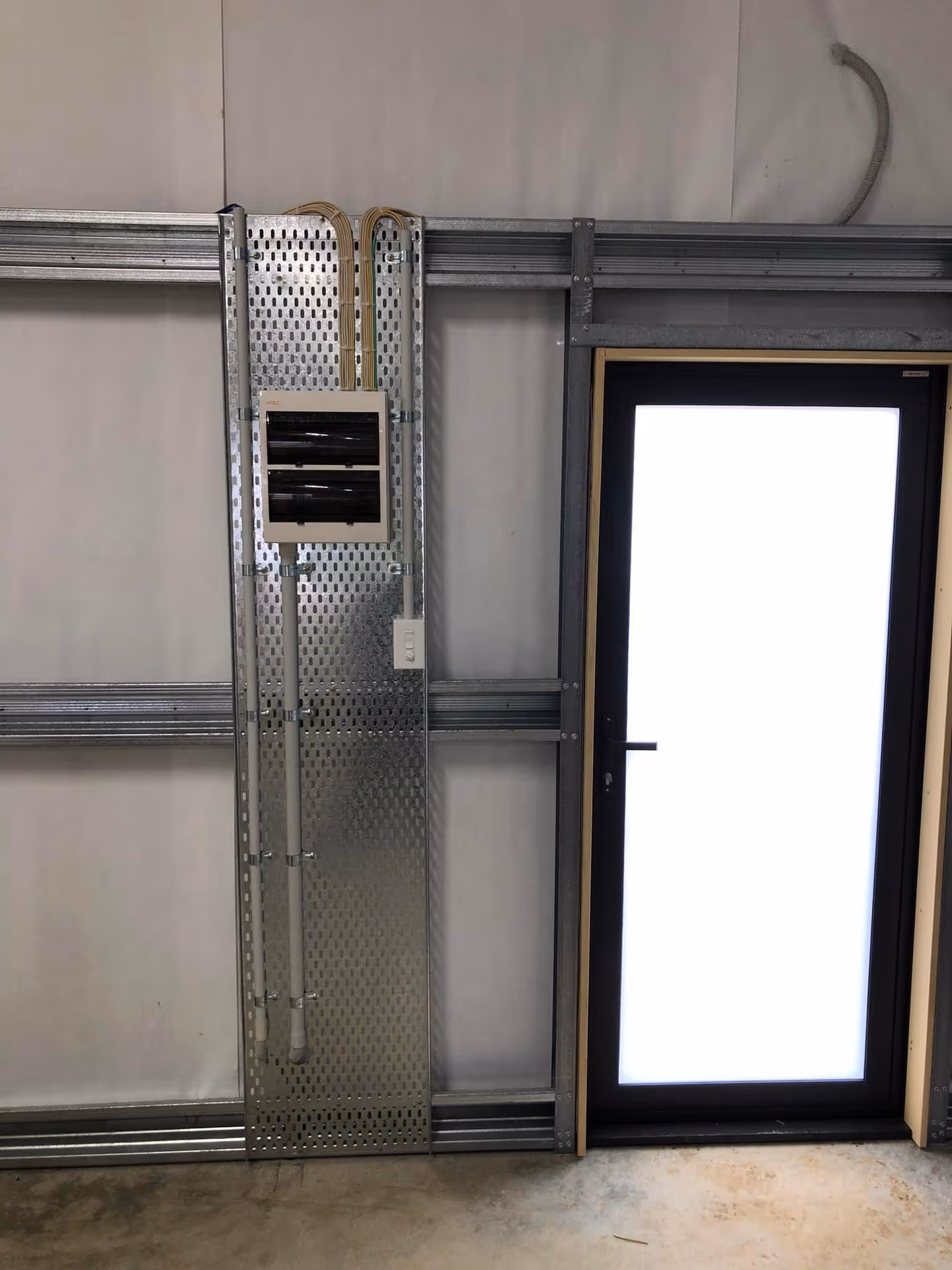 Electrical panel mounted on perforated metal sheet next to a closed door with frosted glass in an industrial wall frame.