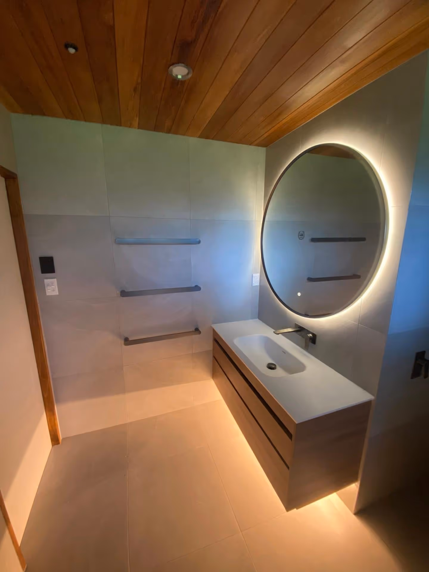 Modern bathroom with wooden ceiling, floating vanity with white sink, backlit round mirror, and ambient floor lighting.