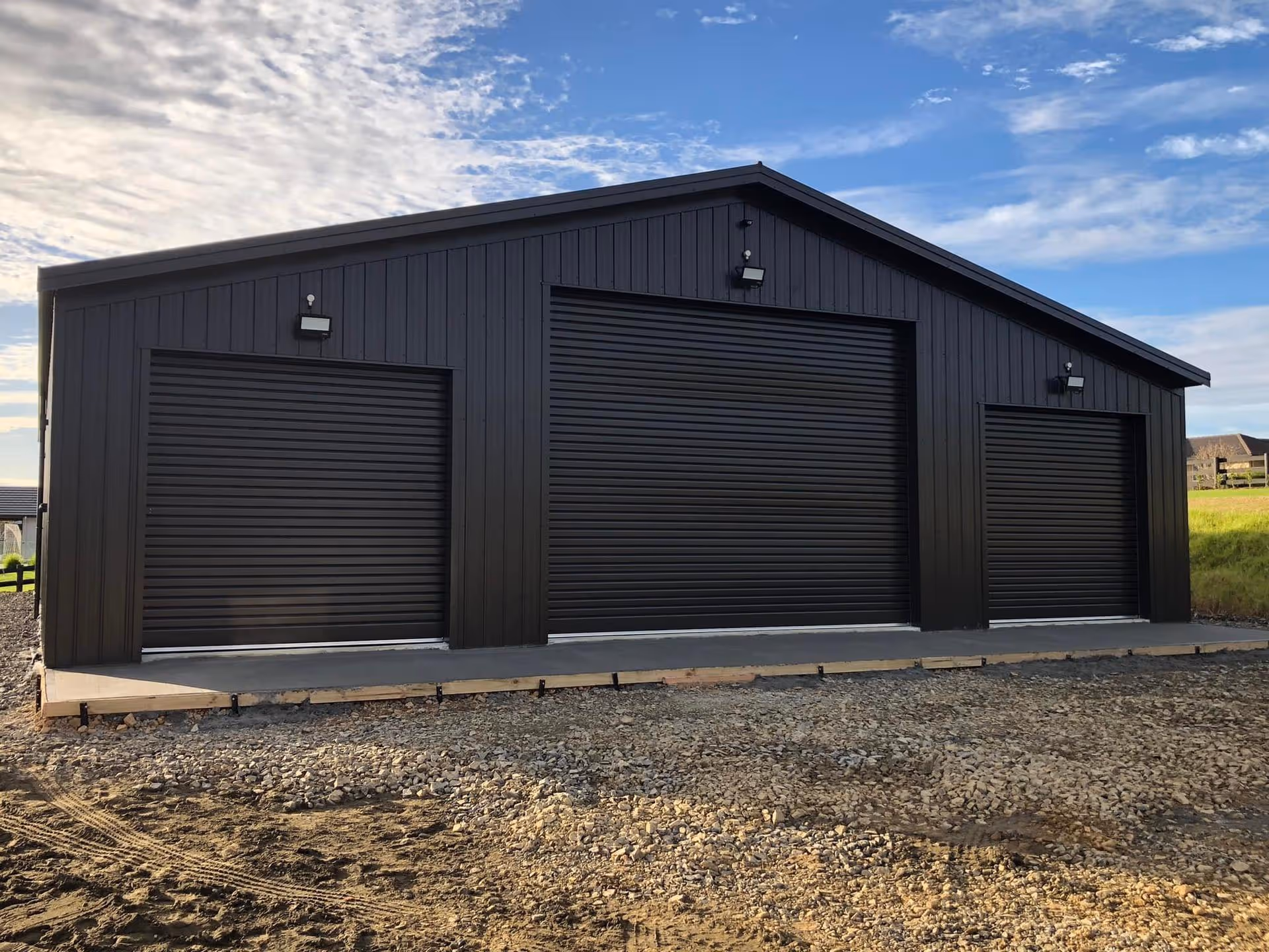 A large black metal storage shed with three closed roller doors, set against a partly cloudy sky and a grassy field, conveys functionality and simplicity.
