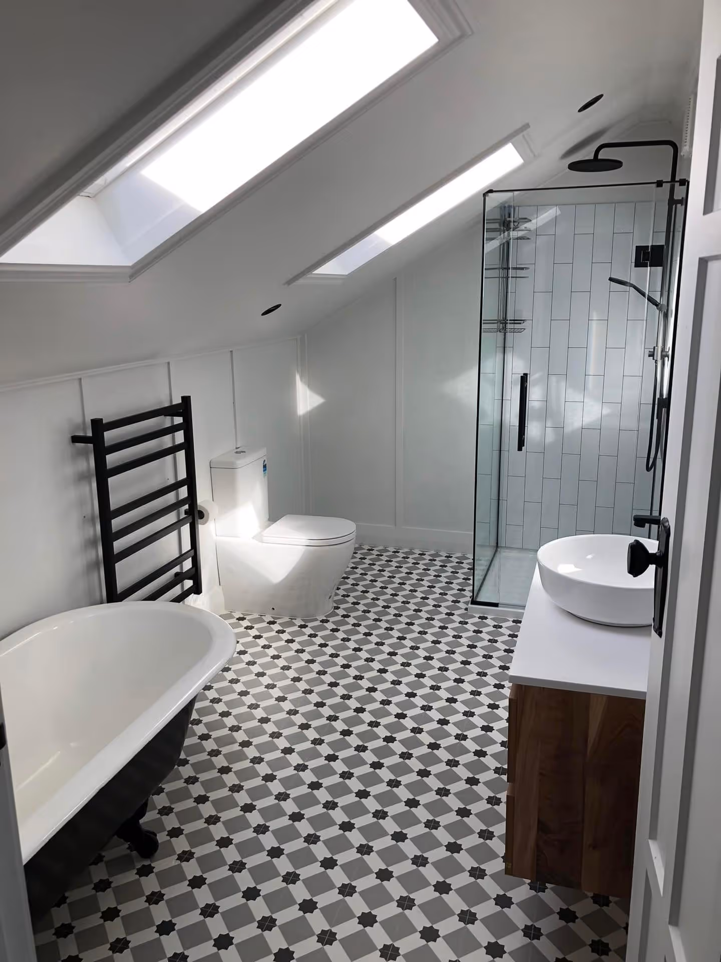 Modern bathroom with sloped ceiling and skylights, featuring a freestanding tub, walk-in shower, patterned tile floor, and white vanity for a sleek ambiance.