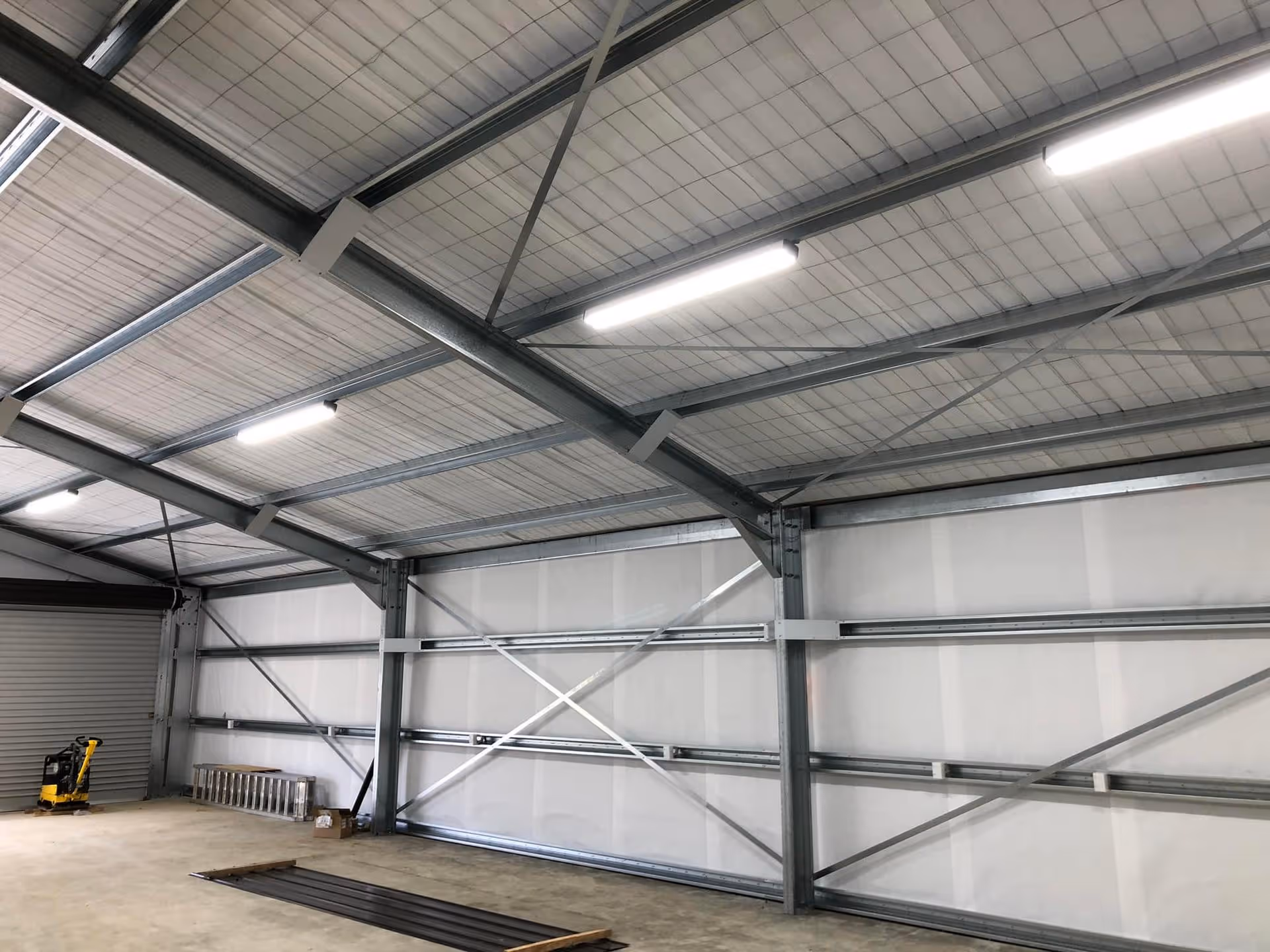 Spacious industrial warehouse interior with a metal framework, concrete floor, and bright overhead lighting. A forklift and ladder are visible near a corrugated door.