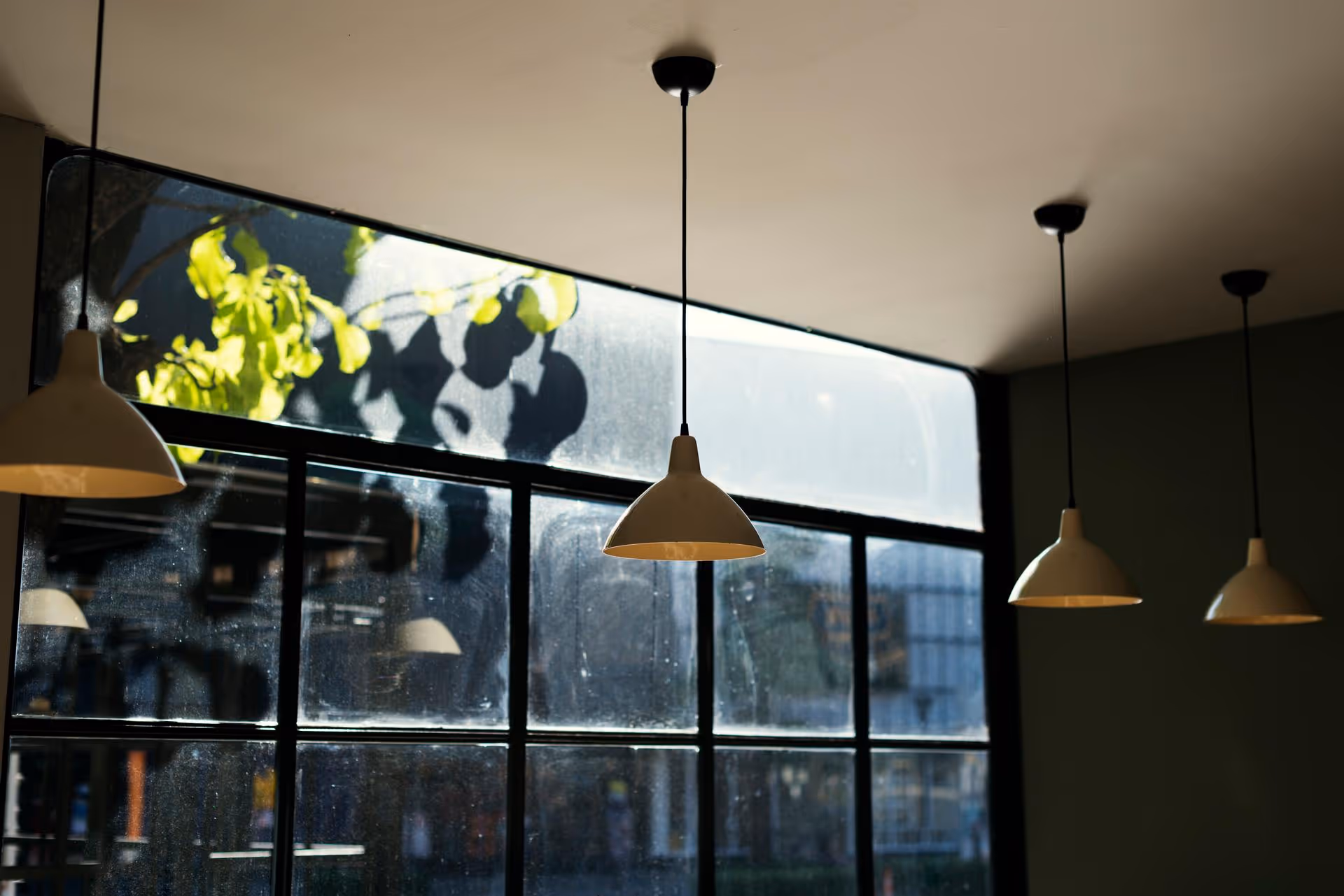Soft sunlight filters through a large window, casting shadows of leaves indoors. Four pendant lights hang from the ceiling, creating a serene and cozy ambiance.
