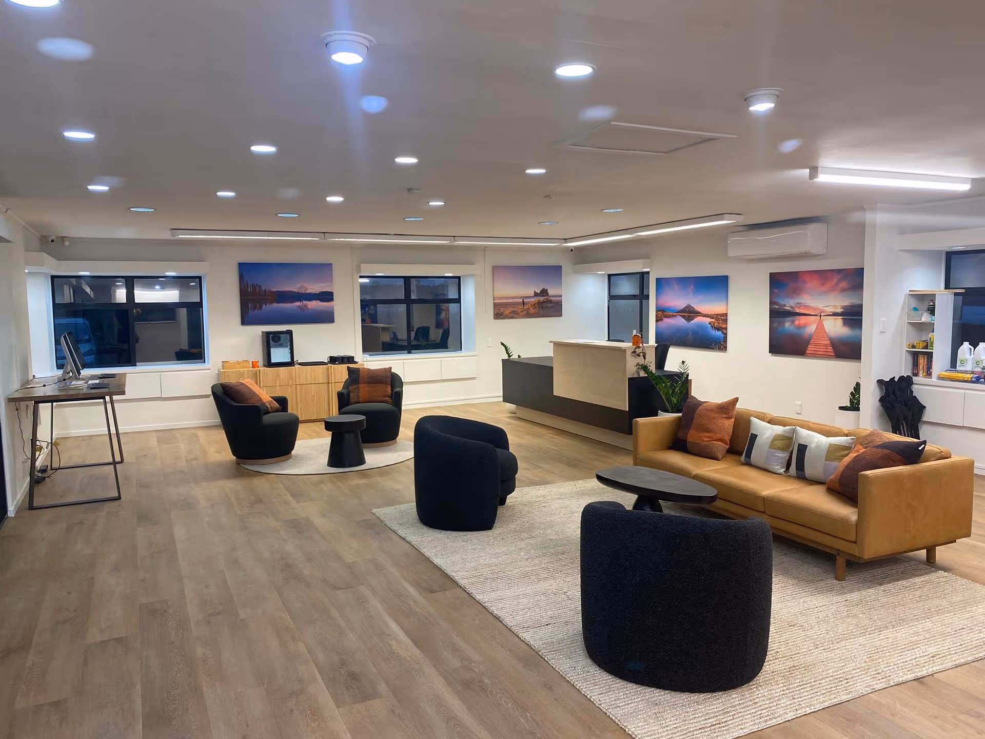 Spacious modern office with wooden floors, mustard sofa, black chairs, and round tables. Large landscape photos adorn white walls, creating a cozy atmosphere.