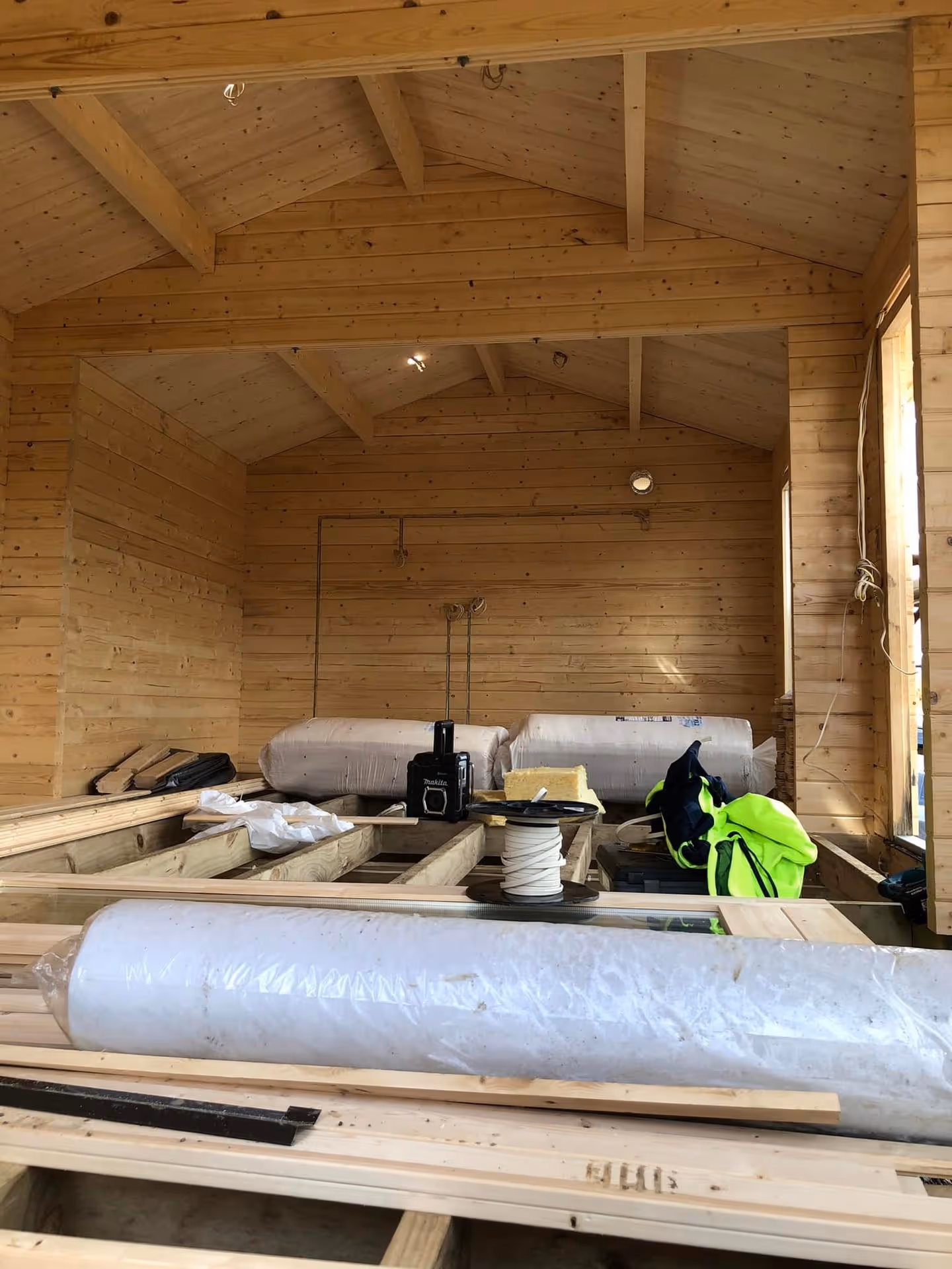 Interior of a wooden cabin under construction, featuring exposed beams and unfinished floors. Construction materials like plastic-wrapped rolls and tools are scattered around.
