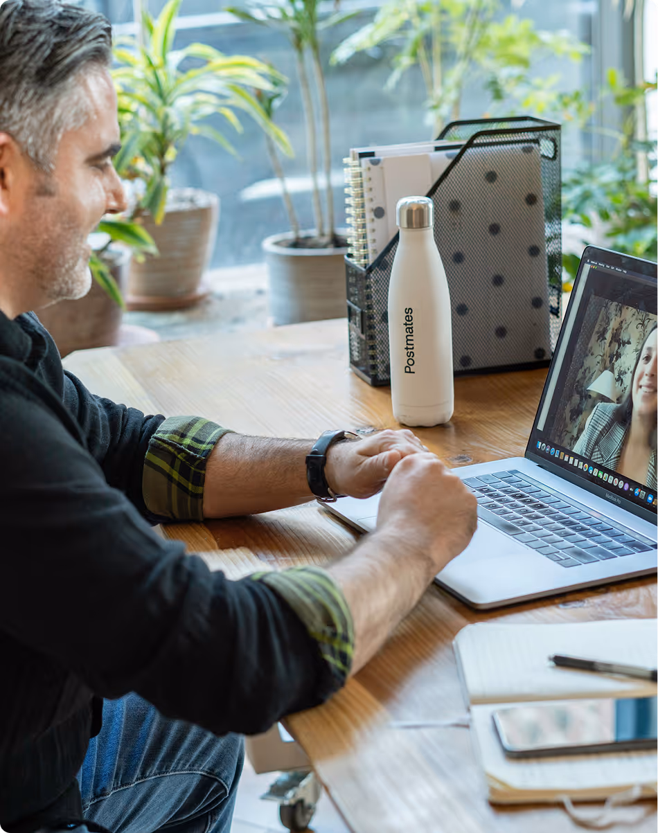 Man in a black shirt with rolled-up sleeves video chatting with a woman on a laptop at a wooden desk with a water bottle and notebook.