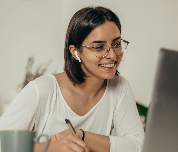 Smiling woman wearing glasses and wireless earbuds, writing with a pen while looking at a laptop screen.