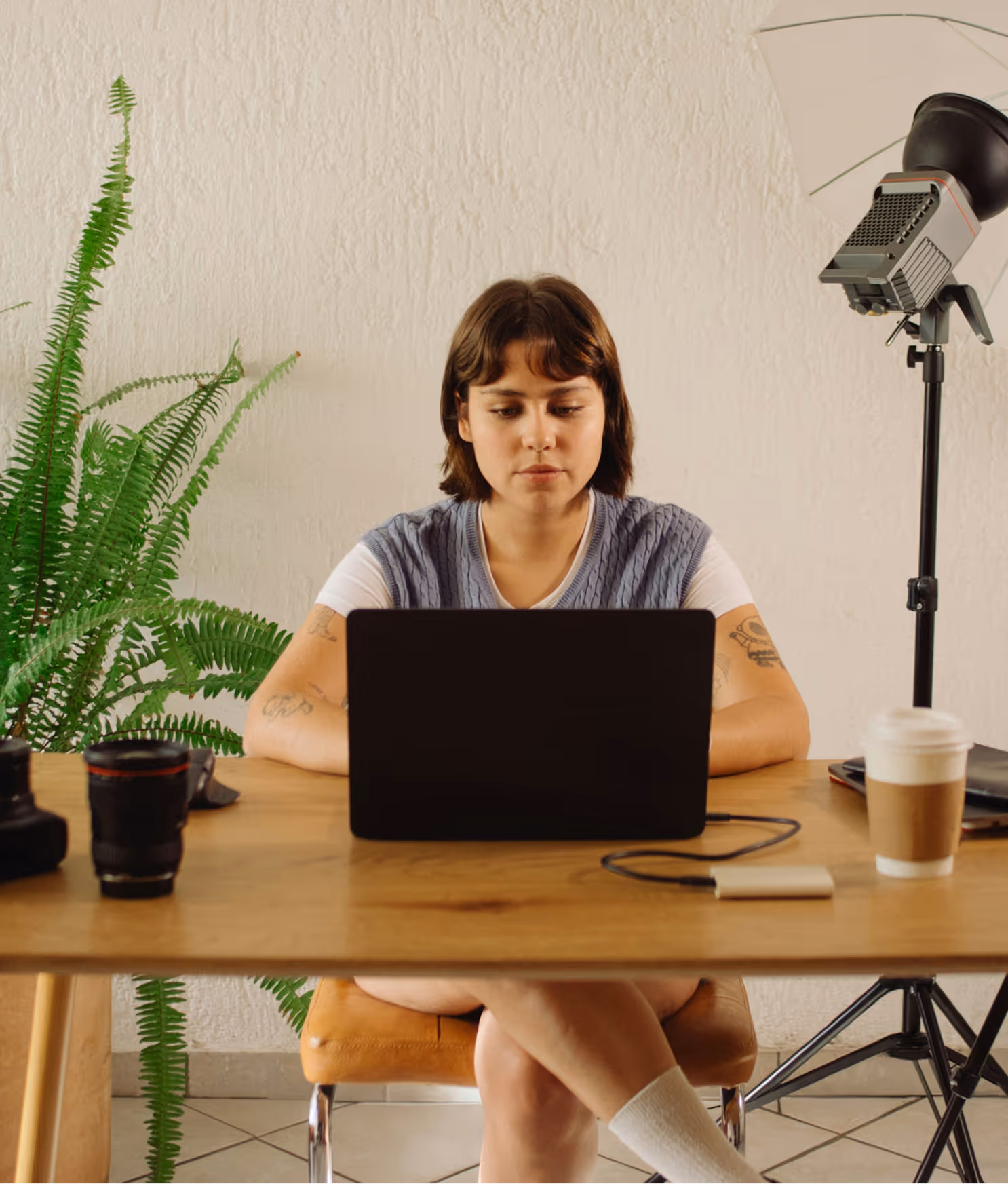 Person with short hair sitting at a desk working on a laptop, surrounded by camera equipment, a plant, and a coffee cup.