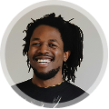 Smiling man with dreadlocks wearing a dark shirt against a light gray circular background.
