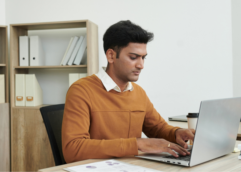 Man in brown sweater working on a laptop at a desk with documents and a coffee cup.