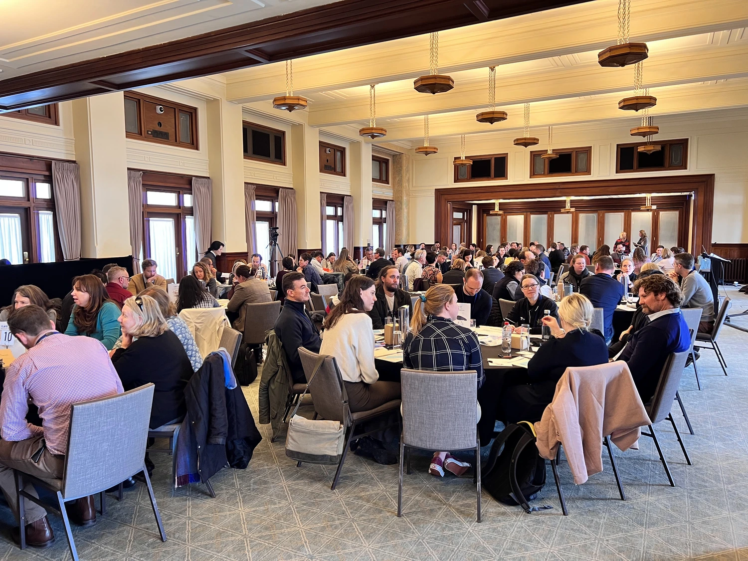 Large group of people seated at round tables in a well-lit conference room engaged in discussion.