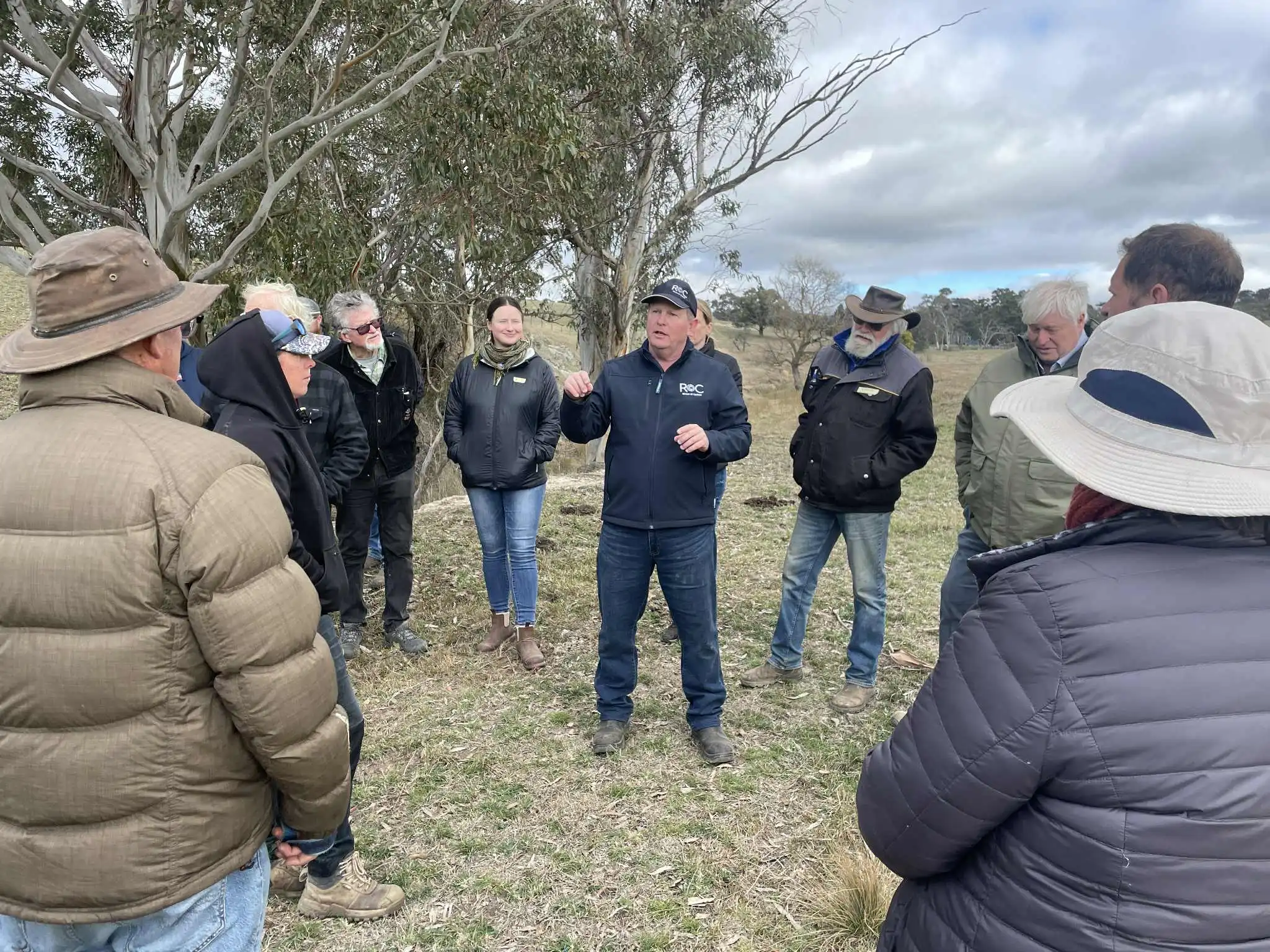 Man in ROC jacket and cap speaking to a group outdoors beside eucalyptus trees on a cloudy day.