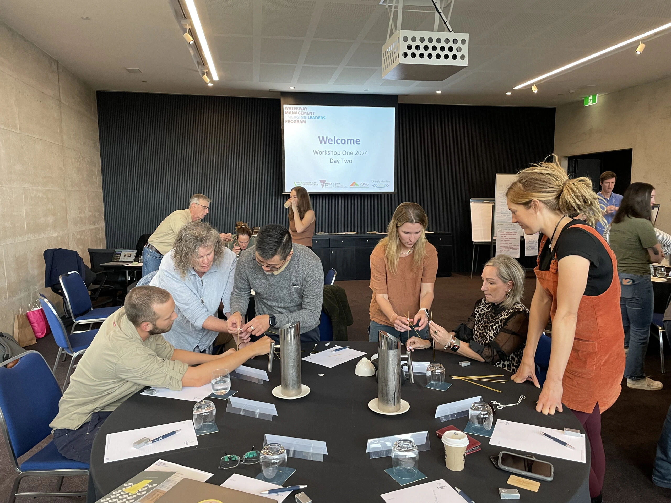 Group of people collaborating around a round table with papers and drinking glasses during a workshop with a 'Welcome Workshop One 2024 Day Two' presentation on screen.