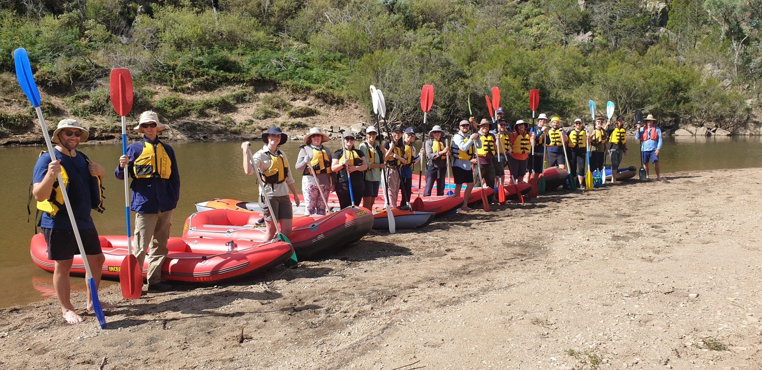 Group of people in yellow life jackets holding paddles standing in a line beside red inflatable rafts on a riverbank.