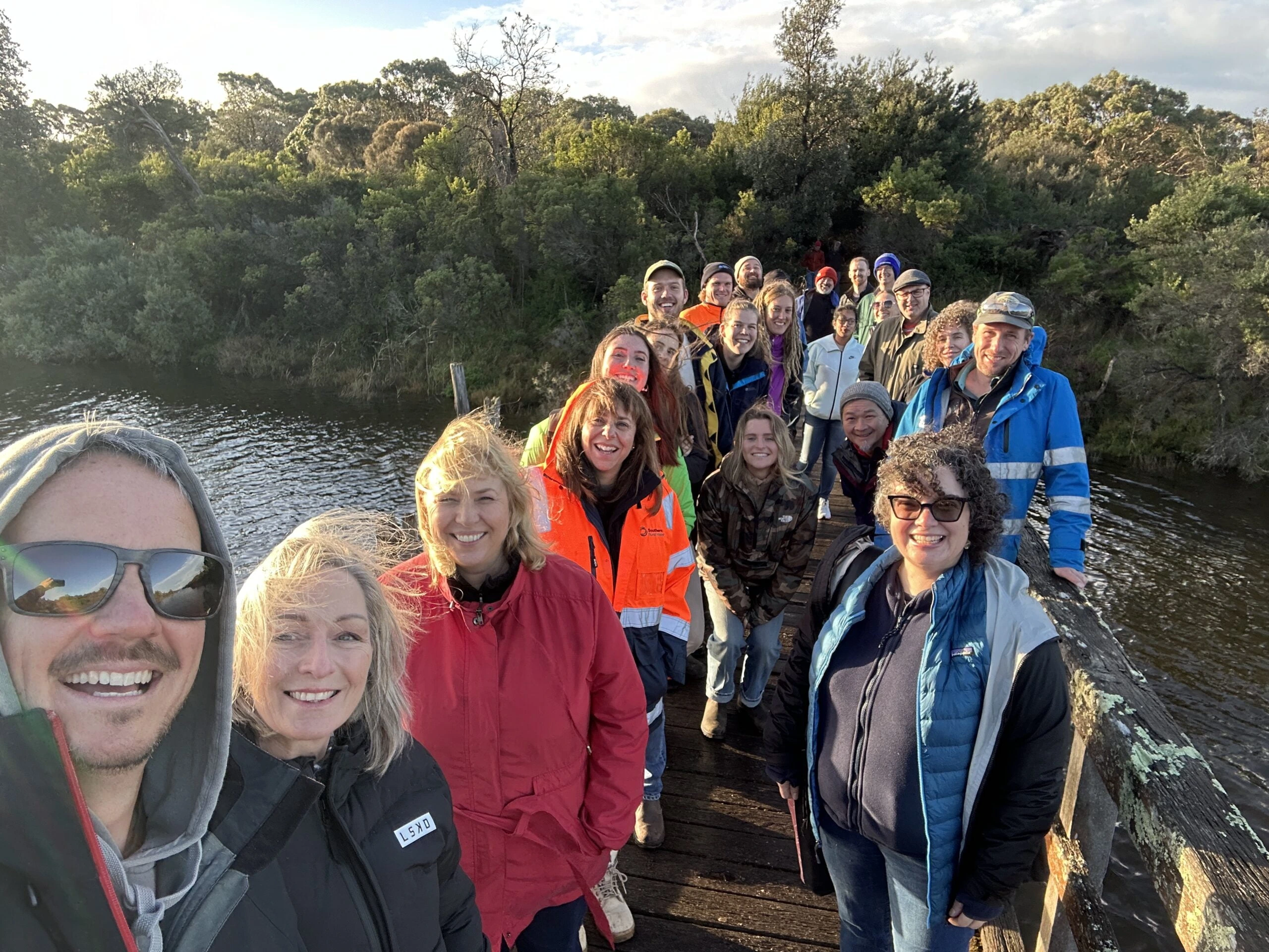Group of people, dressed in jackets and outdoor clothing, standing and smiling on a wooden bridge over water surrounded by dense greenery.