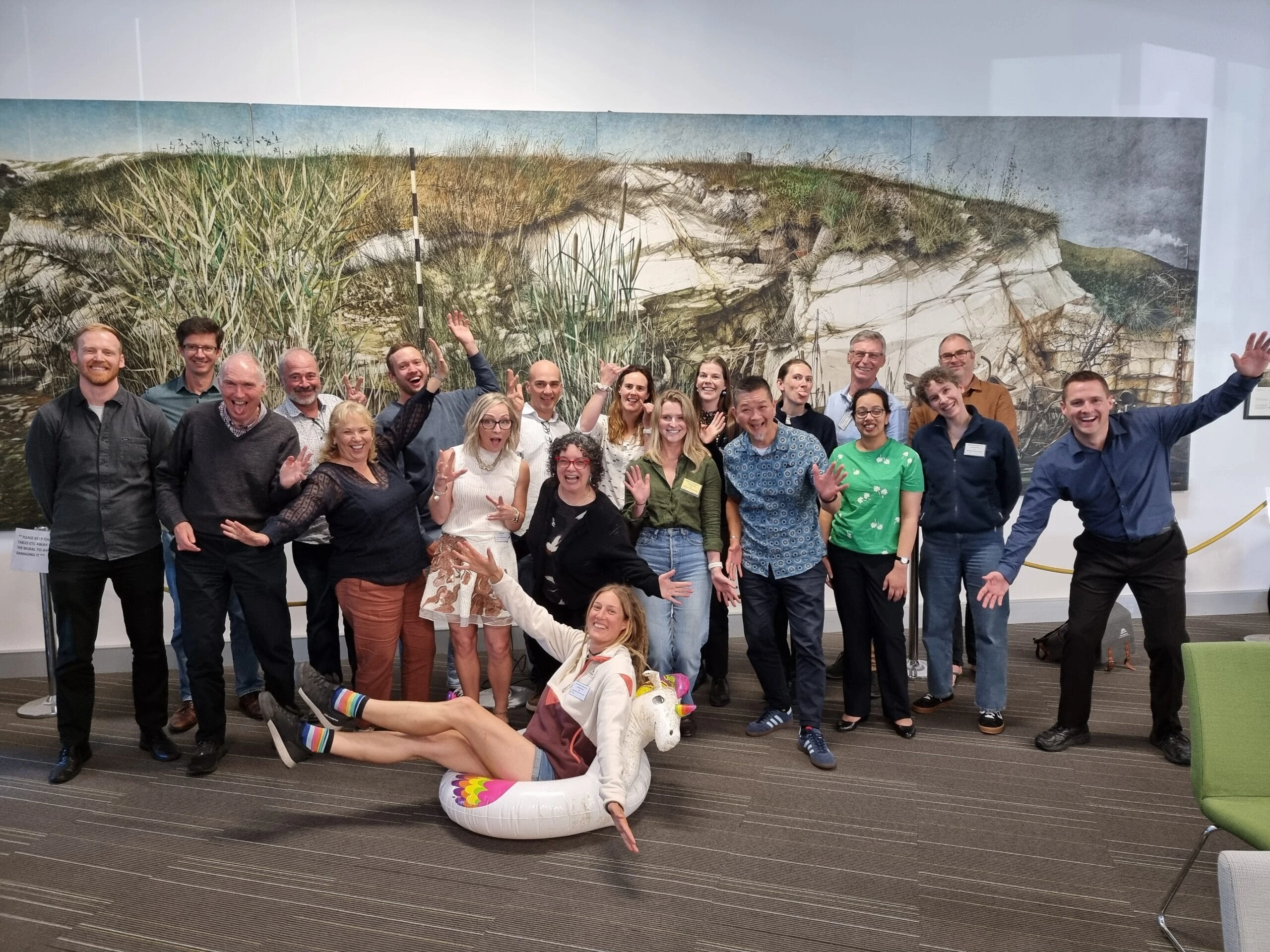 Group of twenty-one people posing indoors in front of a large painting with one person sitting in an inflatable unicorn float, all smiling and making playful gestures.