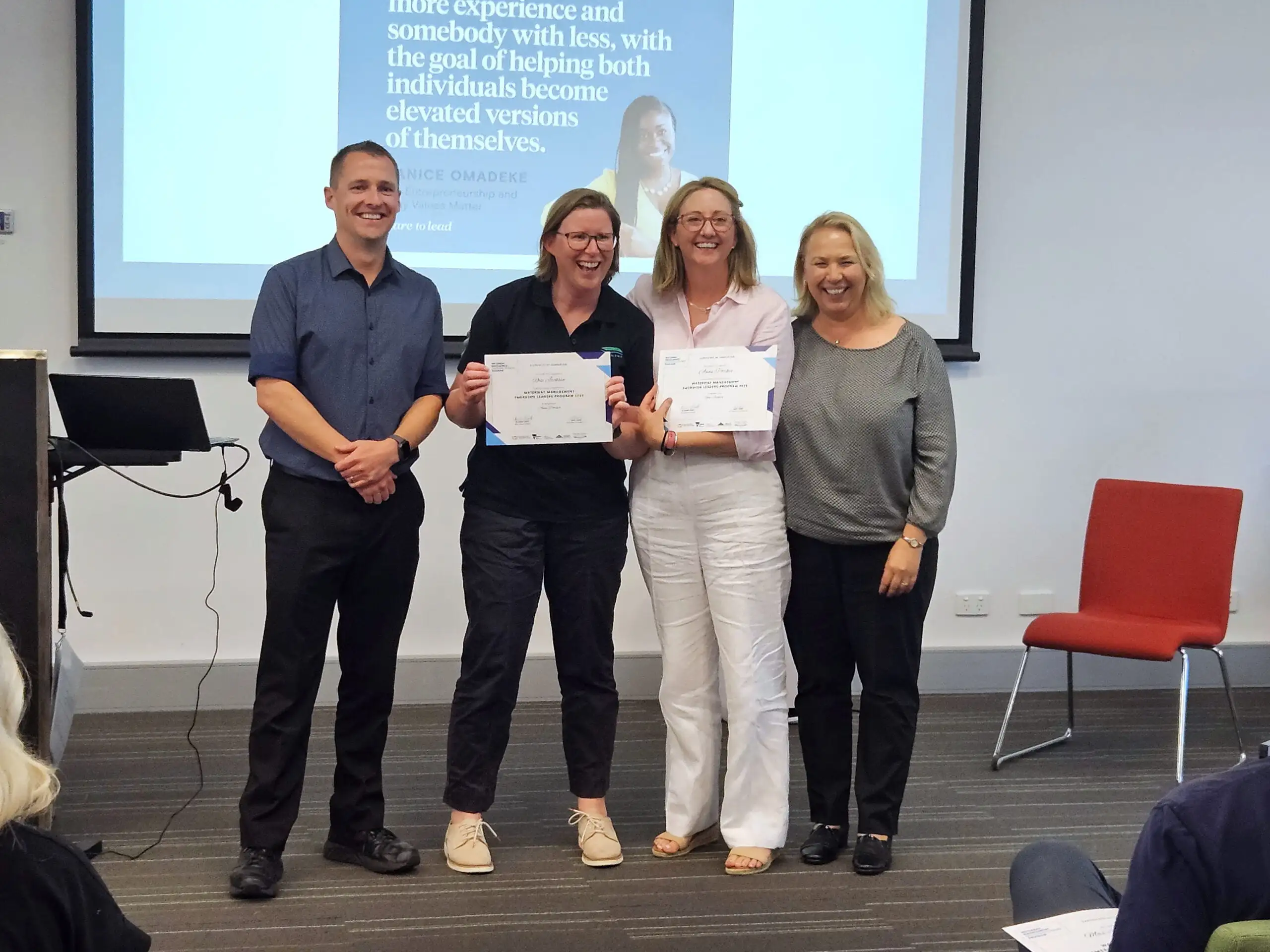 Four adults standing indoors with two women holding certificates, all smiling in front of a presentation screen.