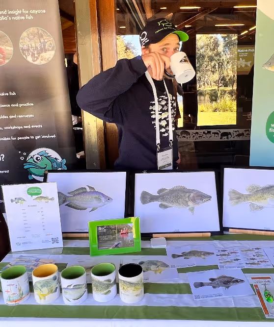 Person wearing a black cap and hoodie drinks from a white mug behind a table displaying fish illustrations and mugs with fish designs.
