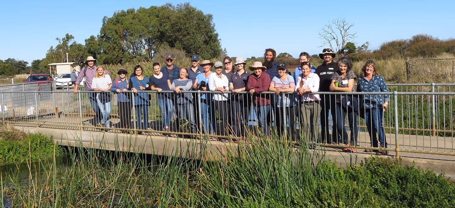 Group of people standing together on a concrete bridge with metal railings over a river, surrounded by green vegetation and trees under a clear blue sky.