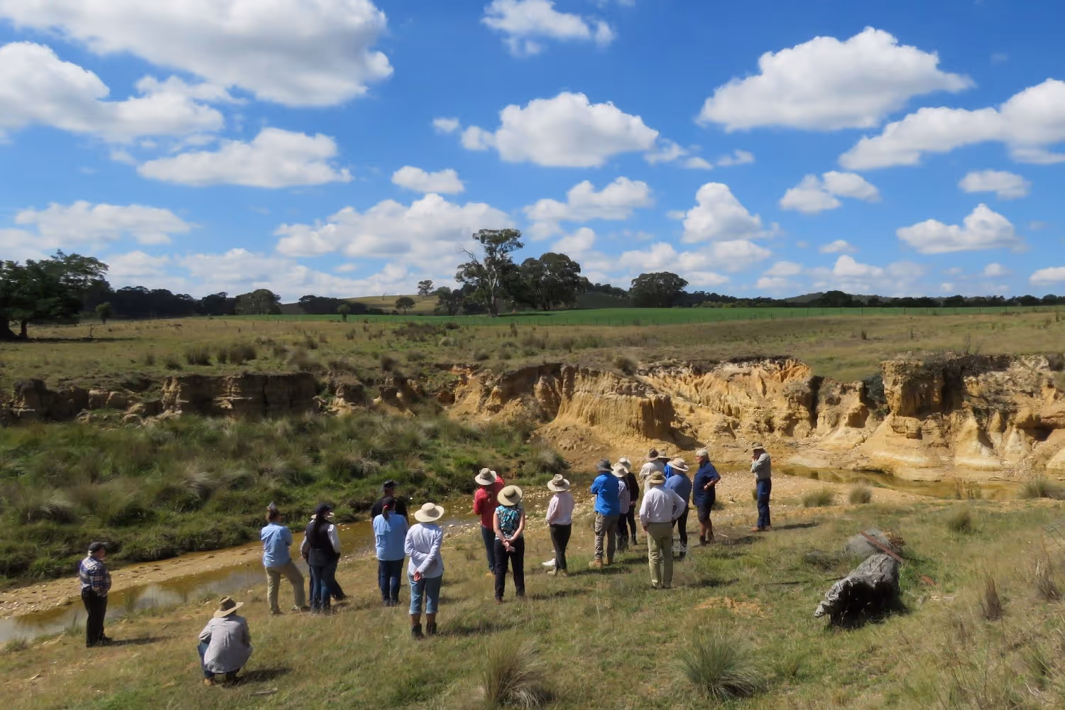 Group of people outdoors near a dry creek bed with eroded soil banks under a blue sky with scattered clouds.
