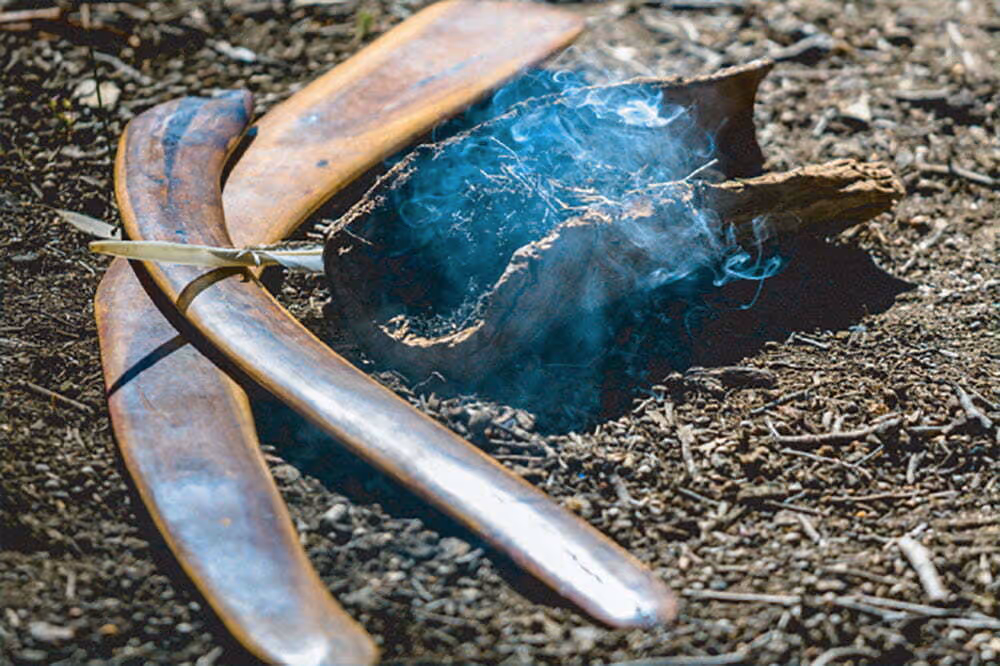 Two wooden boomerangs and a smoking eucalyptus bark fire on dry earth ground.