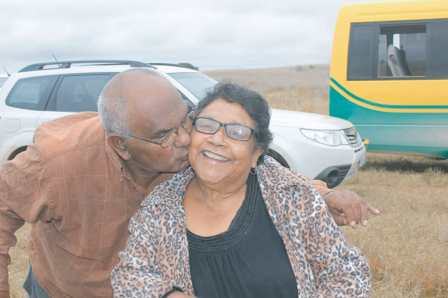 Elderly man kissing an elderly woman on the cheek outdoors with cars and dry grass in the background.